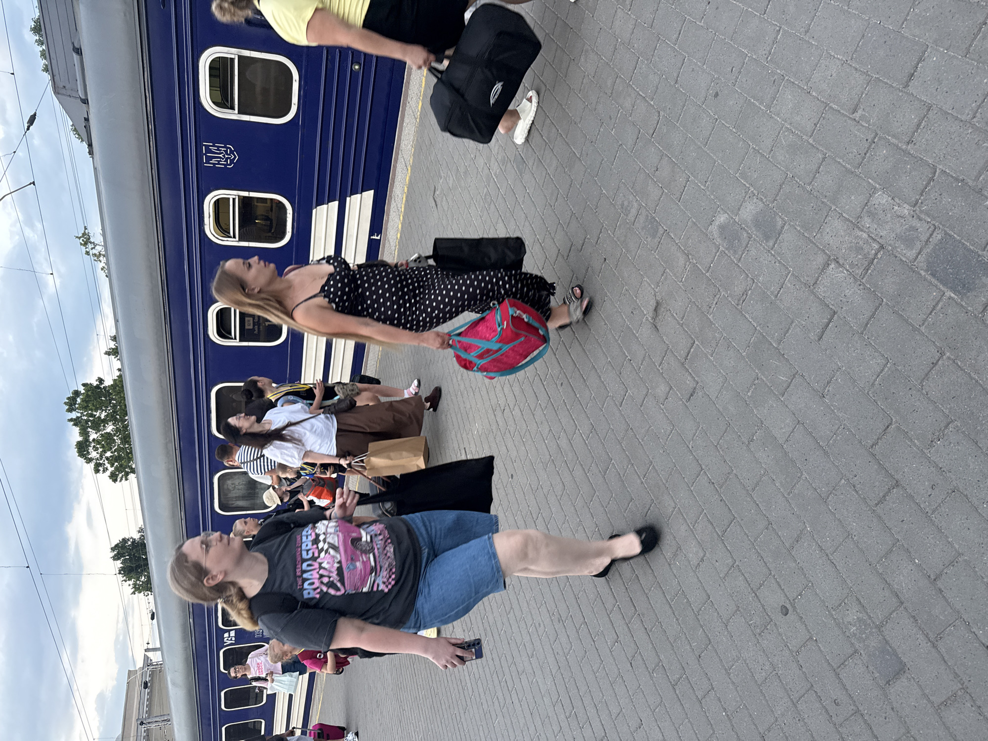 A group of people stand on a paved train platform next to a blue train. The image is taken from a slightly elevated perspective, looking down and to the side. Several individuals are visible, most holding luggage or bags. One woman in the foreground, wearing glasses, a black t-shirt with a graphic design, and denim shorts, is looking upwards. Another woman, further back and to her left, is wearing a black polka dot dress and carrying a bright pink bag. The train cars are blue with white trim and windows. The sky above is cloudy, suggesting overcast weather, possibly late afternoon or early morning. The setting appears to be a train station in Odesa, Ukraine. The overall atmosphere suggests people are waiting to board or disembark a train. Visible text on the woman's t-shirt includes "ROAD SPEED" and other stylized graphics. A Ukrainian coat of arms is visible on the train car.