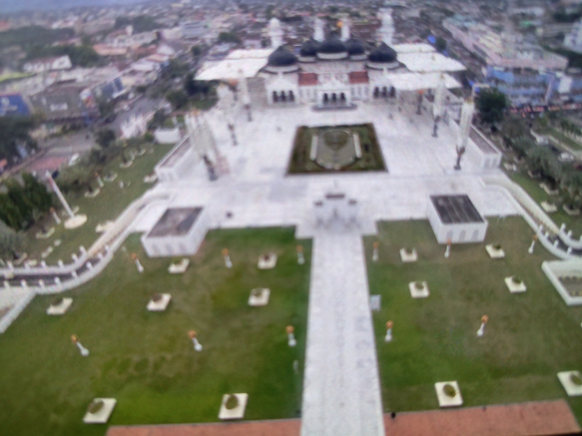 An aerial view of the Baiturrahman Grand Mosque in Gampong Blang, Banda Aceh, Indonesia. The mosque is a large, white building with multiple black domes and a prominent central dome. It is surrounded by a large, open plaza with paved walkways and decorative landscaping. Small, square planters are arranged in rows on the grassed areas. In the distance, the city of Banda Aceh can be seen, with a mix of residential and commercial buildings. The sky is overcast.