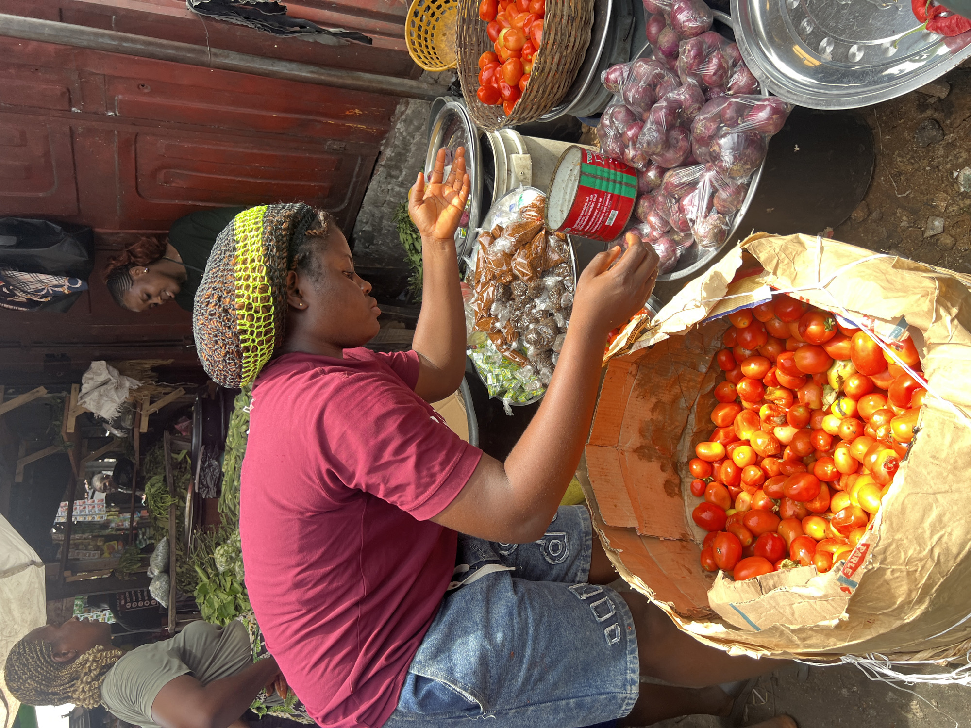 An eye-level, medium shot shows a seated African woman with raised hands. She is in an outdoor market setting in Nkpor, Nigeria. She has short, braided hair covered with a yellow and brown net, wears a maroon t-shirt and denim shorts, and is seated near a large brown paper bag filled with tomatoes.
Around her are various produce items for sale, including baskets of tomatoes and trays of red onions. The market setting includes dark ground and background elements, suggesting a busy, active environment. Other people are visible in the background, indicating the location is a lively commercial space.