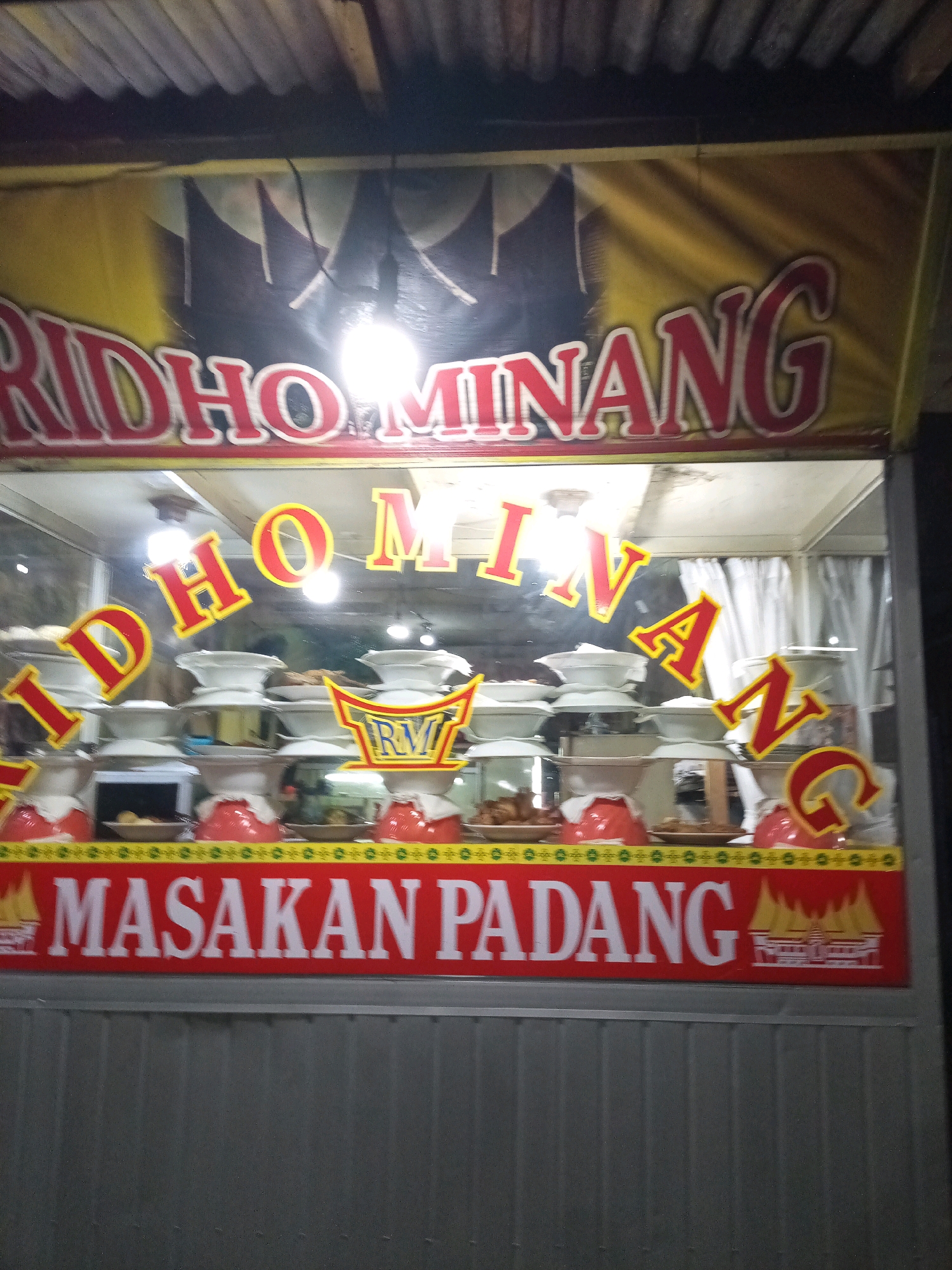 The image captures a food stall in Bekasi, Indonesia, likely a "Masakan Padang" eatery. The stall features a vibrant red banner with white text reading "MASAKAN PADANG." Above this, a larger banner displays the name "RIDHO MINANG" in red letters with white outlines against a yellow and dark blue background. The stall's window displays various dishes of Indonesian cuisine, typically served at Masakan Padang restaurants, arranged in stacked white bowls. Some of the dishes are visible, including what appears to be fried chicken and other savory items. The stall is illuminated by overhead lights, creating a warm glow. The exterior of the stall is simple, with a corrugated metal base.