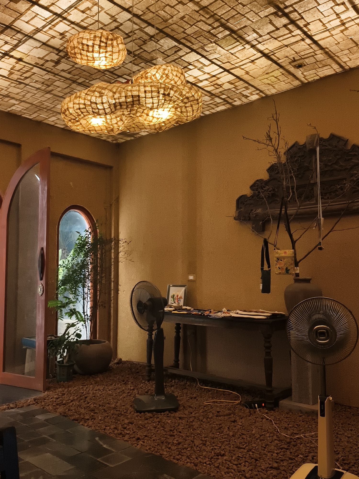 The image depicts an indoor scene, possibly a cafe or restaurant, with a distinctly rustic and natural aesthetic. The ceiling is adorned with a grid-like pattern, illuminated by three large, irregularly shaped pendant lamps made of what appears to be woven natural material. These lamps cast a warm, diffused glow, contributing to the overall ambiance.
The walls are a textured, earthy brown, providing a backdrop for several decorative elements. On the right side of the image, a dark, intricate carved wooden panel is mounted on the wall. In front of this panel, a bare branch with sparse leaves is placed in a large, dark vase, adorned with two small bags hanging from its twigs.
The floor is a combination of dark, rectangular tiles in the foreground and a layer of reddish-brown gravel covering most of the floor area. Two black oscillating fans are present; one is a traditional pedestal fan to the left of a dark wooden table, and another is a more modern, sleek fan on the right. The table is set with various items, including papers, a small framed picture, and what seem to be office supplies.
To the left, an arched doorway, partially ajar, reveals another archway beyond, through which glimpses of greenery from an indoor plant can be seen. A large, round planter sits near this doorway. The lighting suggests an interior space with artificial illumination, possibly supplemented by natural light from the exterior visible through the doorways. The overall impression is one of a thoughtfully designed space aiming for a natural, handcrafted, and slightly bohemian feel. The location in Hoa Lư Ward, Vietnam, suggests a potential Vietnamese influence on the decor and materials used.