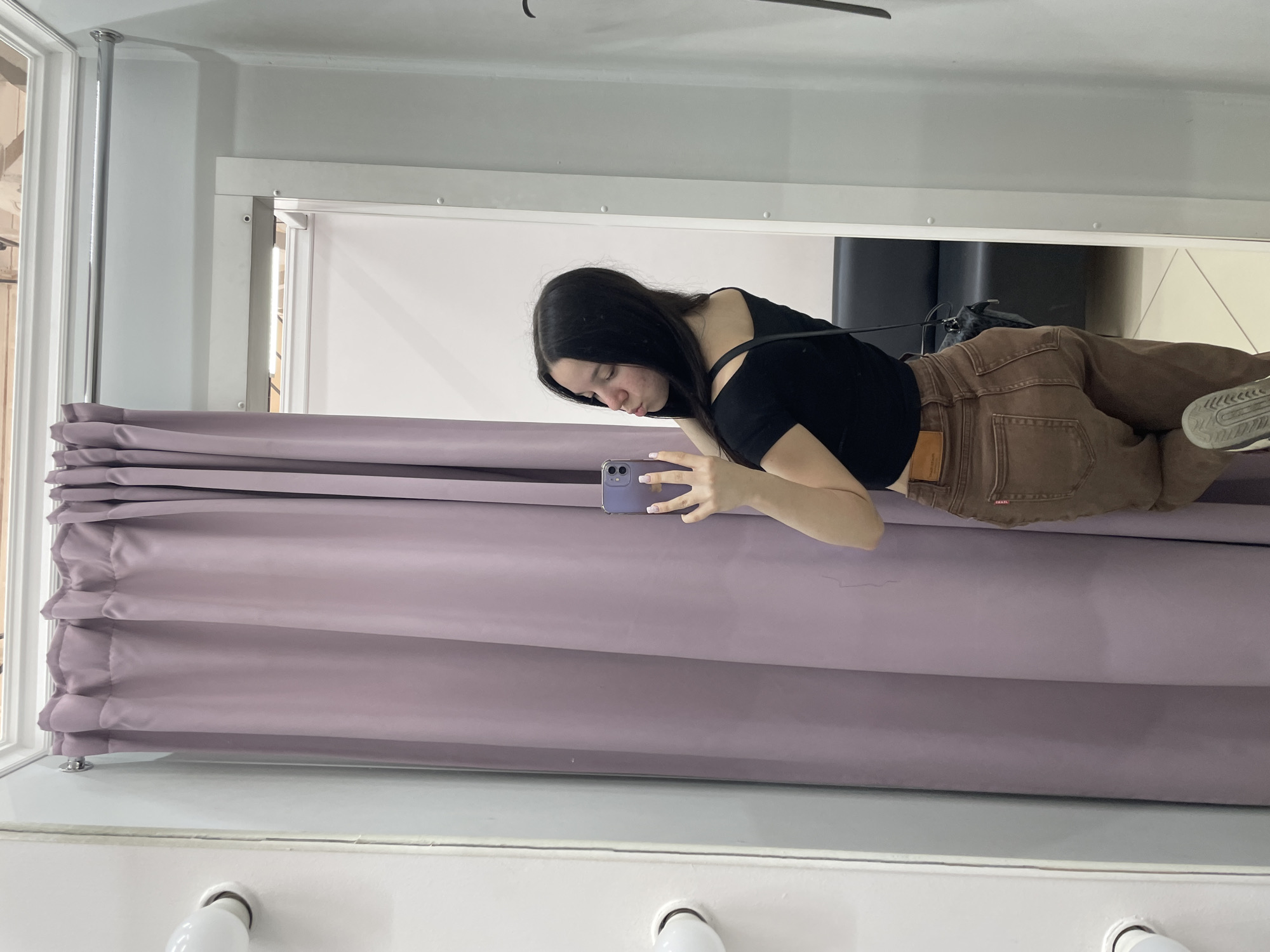 A young woman with long, dark hair is taking a mirror selfie in a dimly lit room, possibly a fitting room. She is wearing a black off-the-shoulder top and brown jeans. Her phone is held up to take the picture, and she is puckering her lips. In the background, a light purple curtain hangs to the left, and a dark couch is visible on the right. The location is identified as Chaikovsky, Russia.