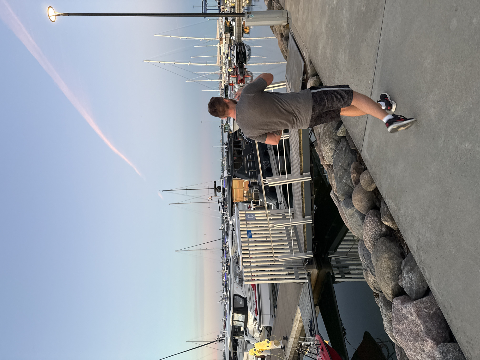 A man wearing a grey t-shirt and dark patterned shorts is walking on a concrete pathway beside a marina. The man is facing away from the camera, and his right arm is bent, with his hand near his face as if holding a phone or other object. He is wearing black and white athletic shoes with red accents. The marina is filled with various boats, including sailboats and motorboats, docked alongside wooden piers. The water is calm and reflects the boats and the sky. In the background, there are more boats and buildings. The sky is a pale blue with a pinkish streak, suggesting it is either sunrise or sunset. A lamppost is visible to the right of the frame. The scene appears to be in a developed waterfront area, possibly a harbor or yacht club. Based on the architecture and vessel types, this scene is likely in Tallinn, Estonia.