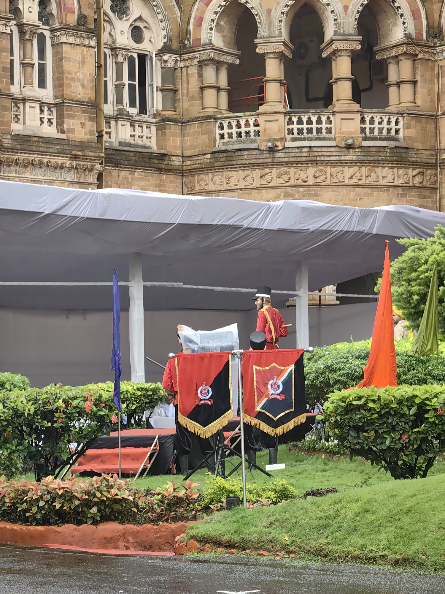 This image captures a ceremonial scene outside the Chhatrapati Shivaji Maharaj Terminus in Mumbai, India. In the foreground, two large black and red flags with a crest are displayed. Behind the flags, a person in a red ceremonial uniform with a black hat stands on a raised platform, holding a baton. Another person in a similar uniform is partially visible to the right. The background features the iconic Victorian Gothic architecture of the railway station, with its ornate stone carvings and arched windows. A white tent or canopy is erected over the platform area, suggesting an outdoor event. Lush green bushes and grass surround the platform, indicating a landscaped area within the city. The overall atmosphere appears formal and possibly part of a parade or official function.