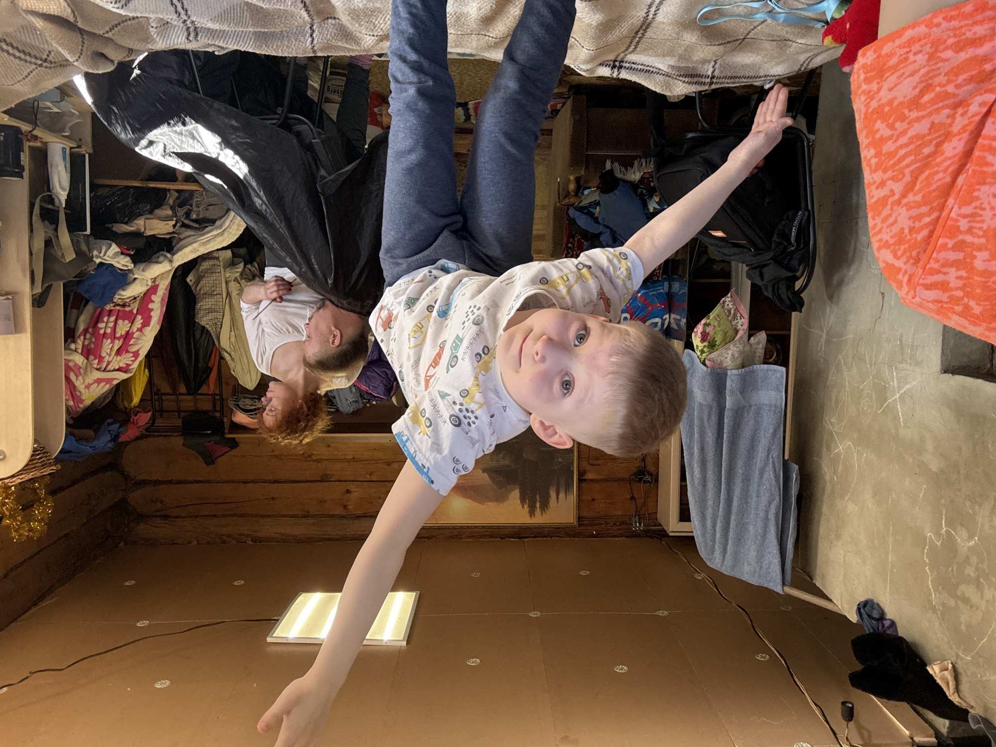 A young boy wearing blue pants and a white t-shirt printed with vehicles is hanging upside down from a ceiling in a room in Nizhnyaya Chermoda, Russia. His arms are outstretched, one reaching towards a square light fixture on the floor and the other towards an orange towel. In the background, an adult woman with reddish hair is hanging upside down near a dark plastic sheet and clothes hanging from a closet. The walls of the room are made of logs. The floor is brown. A gray towel hangs nearby. The picture is taken from the perspective of someone standing on the floor looking up. The entire scene is upside down.