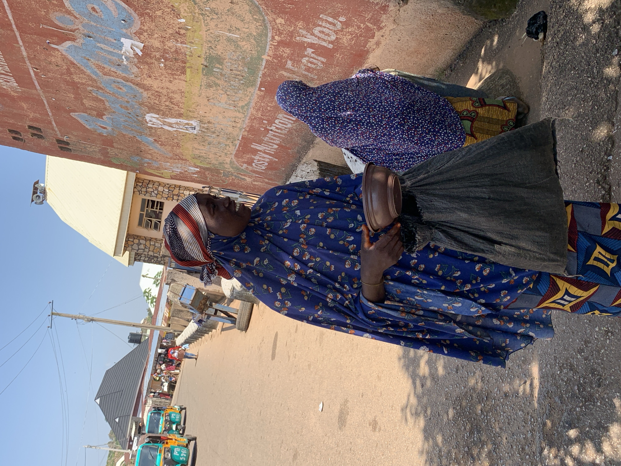 A woman stands in the foreground of the image, wearing a blue hijab with floral patterns and a colorful patterned skirt. She is holding a small brown bowl in her left hand and a dark bag in her right. She is looking to her left with a slight smile. Behind her, another person also wearing a hijab is visible. They are standing on a dirt road in what appears to be a marketplace or street in Jos, Nigeria. In the background, there are shops, vehicles (including several bright yellow and blue tricycles, also known as "Keke Napep"), and buildings. A large red wall with faded writing is on the right side of the image. The sky is clear and blue, and the sun is shining. The ground is a mix of dirt and gravel, with some shadows cast by trees and structures.
