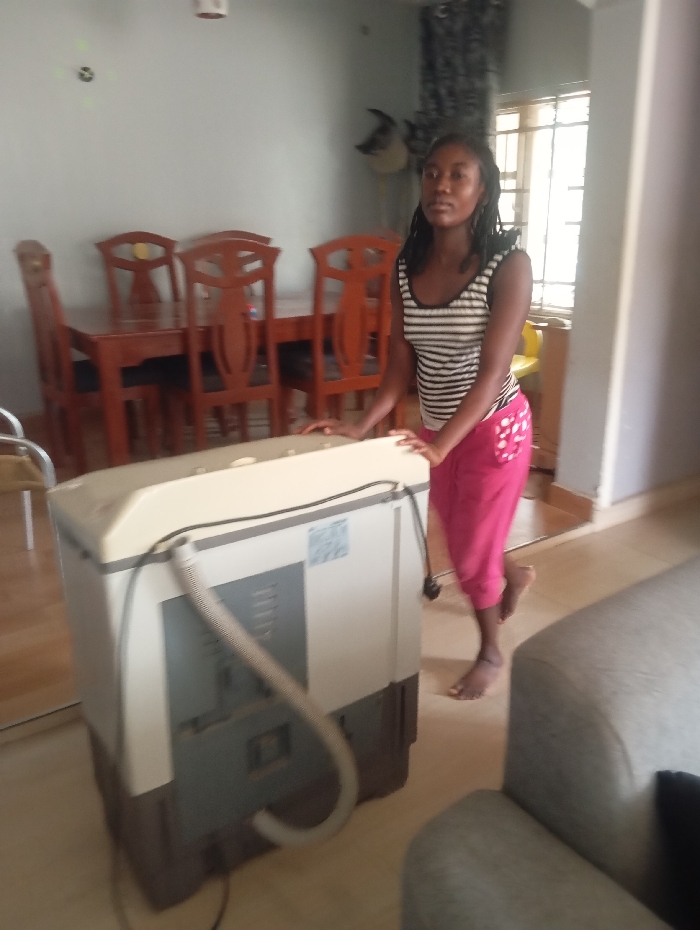 An indoor scene in a domestic residence in Makurdi, Nigeria, depicts a young Black female standing barefoot next to a semi-automatic, top-loading washing machine. The female, with dark hair, wears a black and white striped sleeveless top and pink capri pants with white polka dots on the left pocket. Her hands rest on the cream-colored top surface of the washing machine, which is grey on its lower section and features a visible grey corrugated drain hose and electrical cords. Her body is angled, with her right leg slightly bent and heel raised, and she looks towards the left of the frame. In the background, a wooden dining table surrounded by six wooden chairs with decorative backs is visible against a light grey wall. A window with horizontal security bars and dark patterned curtains is present on the right, illuminating the room. A portion of a light grey upholstered sofa with a dark cushion is in the foreground, to the right. The floor is light-colored.