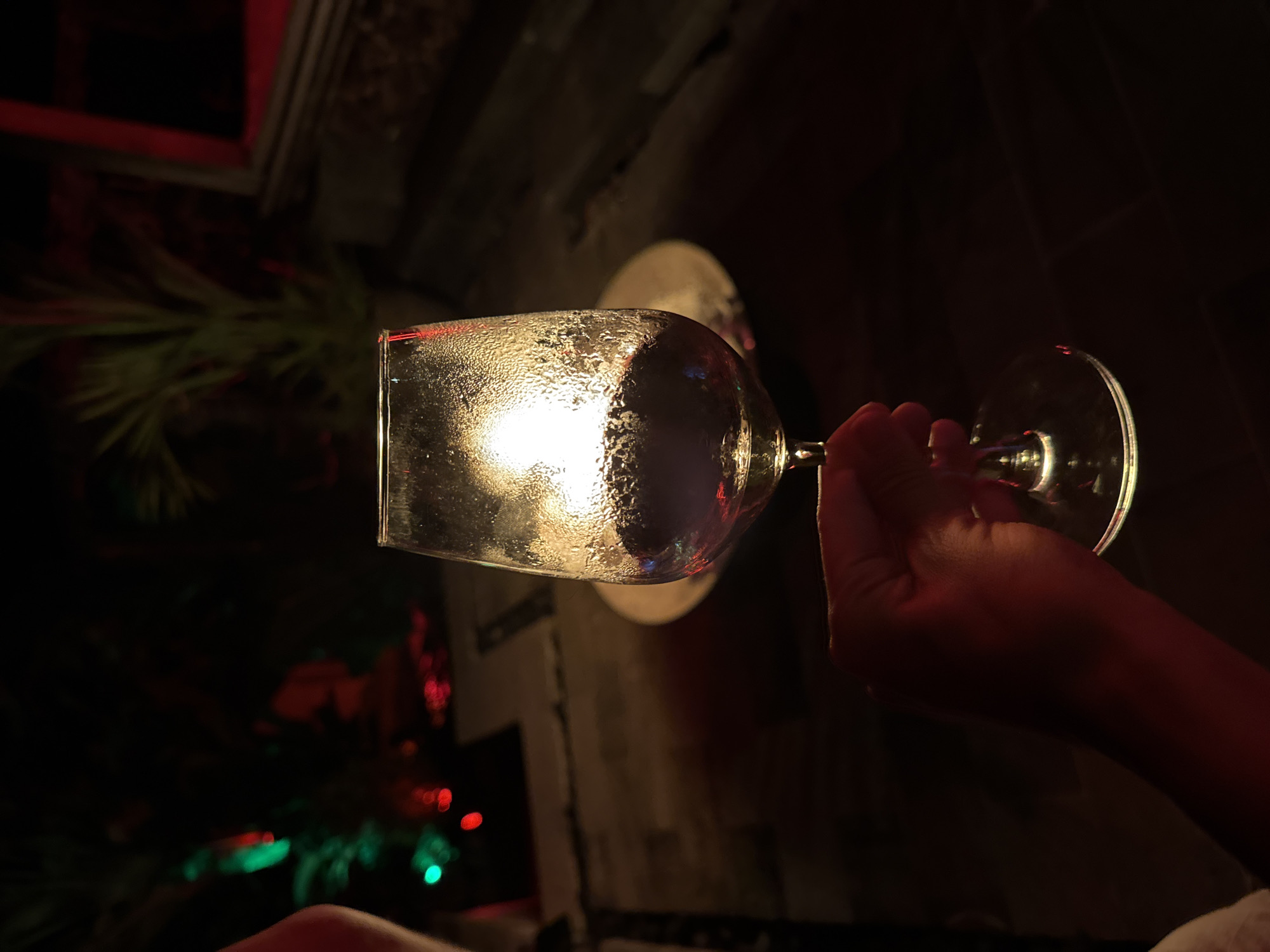 A hand holds a condensation-covered wine glass in a dim, indoor setting in Ungasan, Indonesia. Red and green lights are visible in the background, suggesting a holiday or decorative lighting setup. A white object, possibly a plate or coaster, sits behind the glass. The hand appears to be gripping the stem of the wine glass. The environment is dark, with hints of furniture or architectural elements in the periphery.
