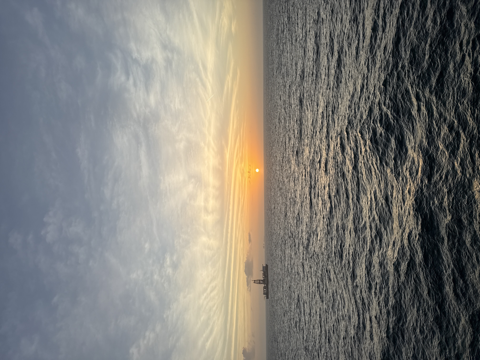 The image shows a sunset over the ocean. The left side of the image is a sky with clouds, with the sun setting. The right side is water with ripples. A rig is visible on the horizon. Location is unknown.