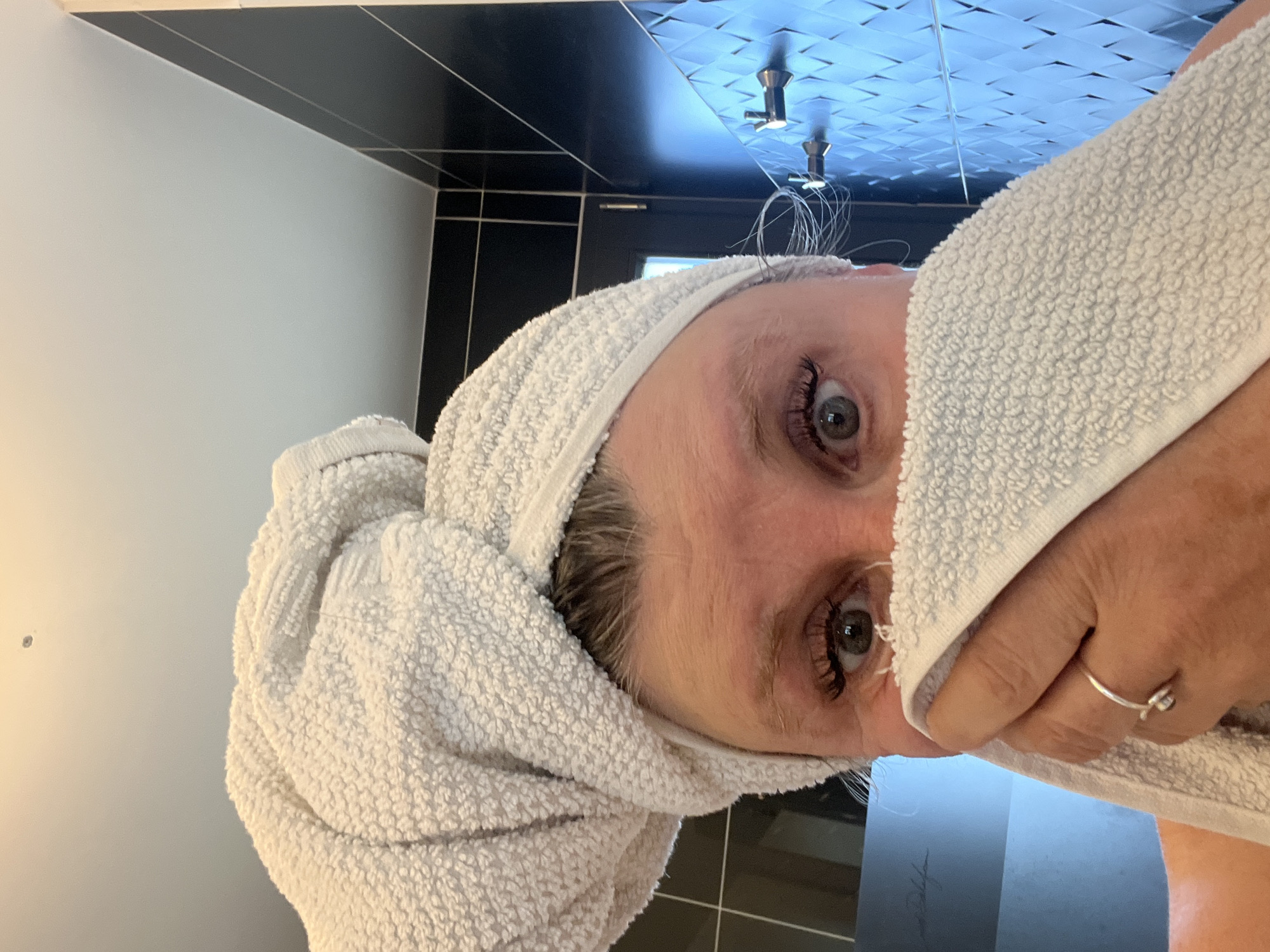 A person, likely a woman, is seen from a close-up, high-angle perspective. She has a white towel wrapped around her head like a turban and is holding part of the towel to her face. Her gray eyes are visible, and she wears a ring on her finger. The background suggests a bathroom setting with black and white tile. The location appears to be a bathroom in a private residence. The city is not relevant.