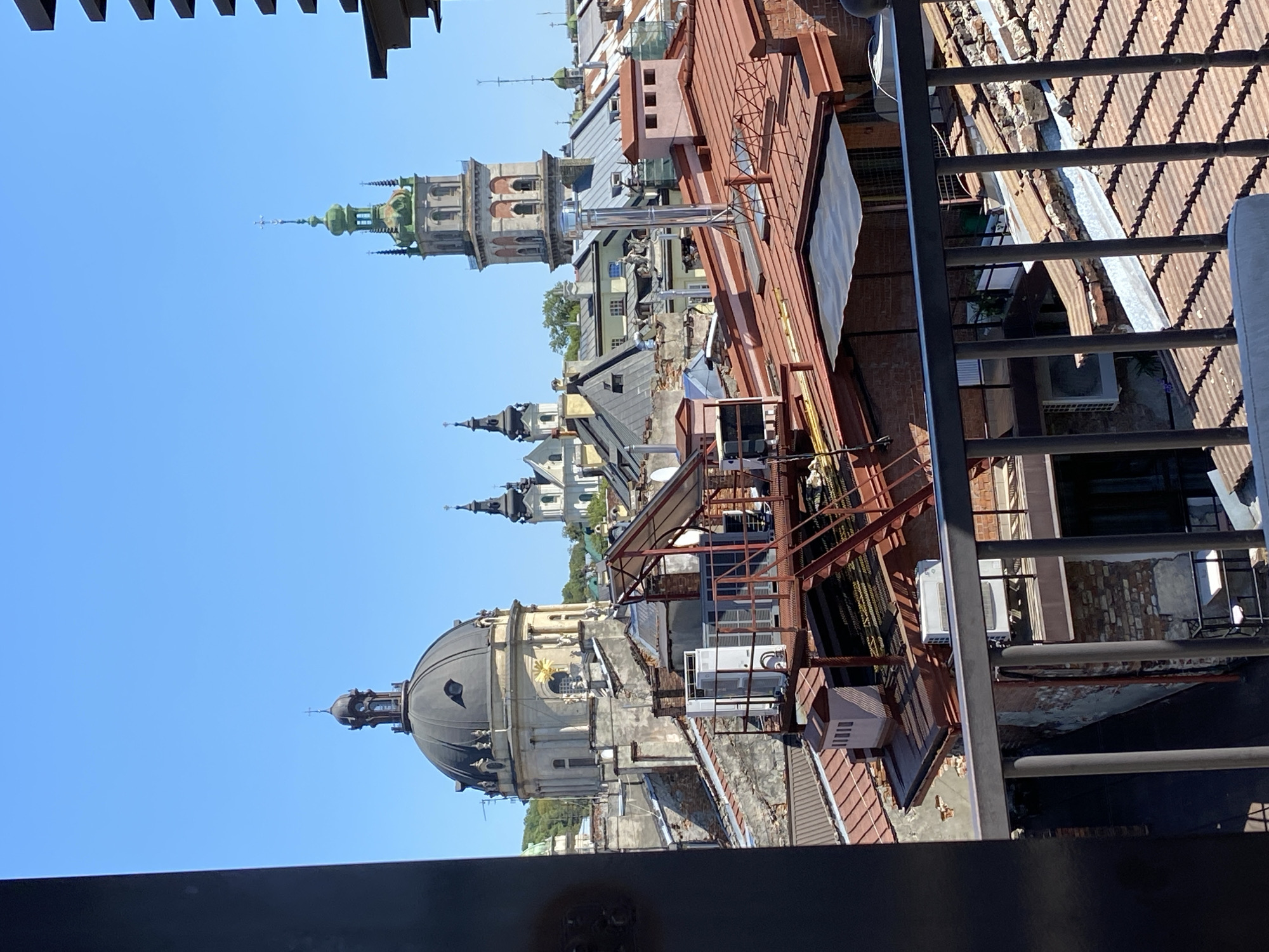The image captures a rooftop view of the historic city of Lviv, Ukraine, under a clear blue sky. Prominently featured is the dome of the Armenian Cathedral of Lviv, a significant architectural landmark. To the right of the dome, the bell tower of the Bernardine Church, also known as the Church of St. Andrew, rises with its distinctive green spire. The foreground is dominated by a network of terracotta tiled roofs, some showing signs of age and wear. Several buildings with weathered facades and exposed brickwork are visible, interspersed with industrial elements like air conditioning units and metal scaffolding. The scene is devoid of any people. The overall impression is one of urban texture and architectural heritage.