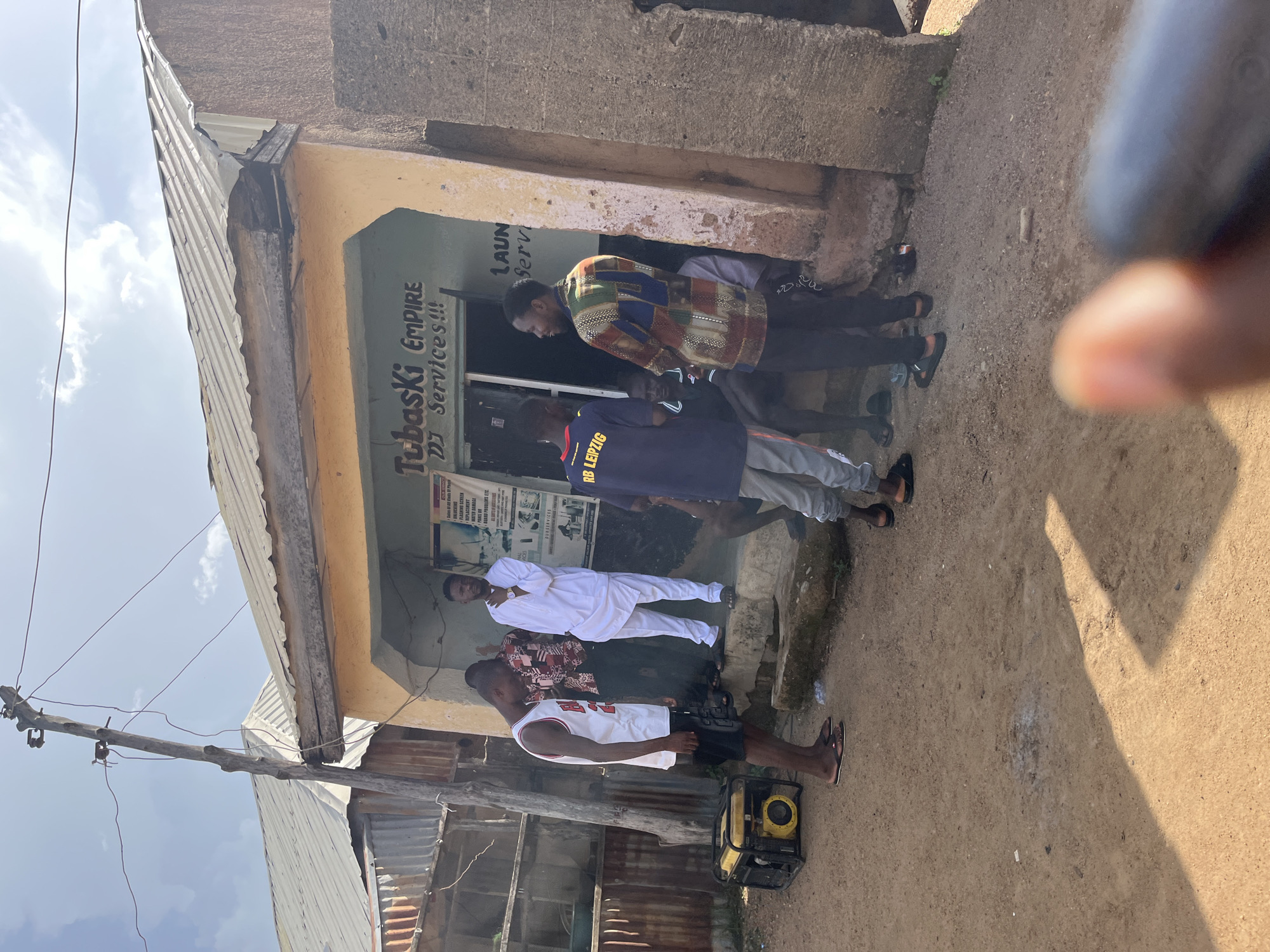 The image depicts a street scene in Jos, Nigeria. In the foreground, a yellow generator is placed on the ground next to a person wearing a white tank top and black shorts. To their right, a group of men are gathered outside a building with a sign that reads "Tubaski Empire DJ Services!!!". One man is wearing a white traditional outfit, while another is in a multicolored shirt. Other individuals are visible in the background, engaged in conversation. The environment appears to be an urban setting with corrugated iron structures and a partially paved ground. The sky is cloudy.