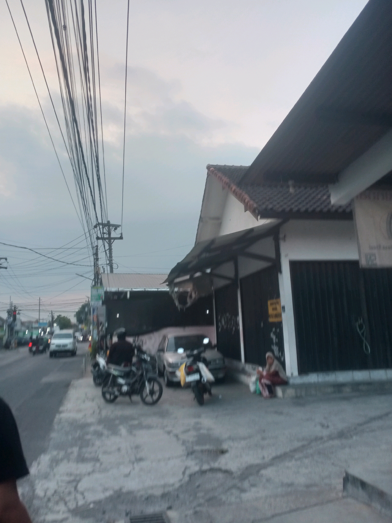 The image shows a street scene in Maguwoharjo, Indonesia, possibly during the early morning or late afternoon, given the soft lighting and overcast sky. A prominent feature of the scene is a dense network of overhead electrical wires, supported by utility poles, spanning across the frame.
On the right side of the street, a building with a dark, corrugated metal roof and a white facade is visible. The building has several entrances covered by dark, rolling metal shutters. A person, likely a woman wearing a headscarf and a maroon and white garment, is sitting on the curb in front of the building, with bags near her. Adjacent to her is a black motorcycle.
On the left side of the street, there is a road with several vehicles, including cars and motorcycles, moving in the distance. A white car and a dark motorcycle are closer to the foreground. The sidewalk in the foreground is made of concrete with visible cracks and some puddles or wet patches, indicating recent rain or high humidity. The overall atmosphere is that of a typical urban street in Southeast Asia, with a mix of commercial buildings, infrastructure, and daily life.