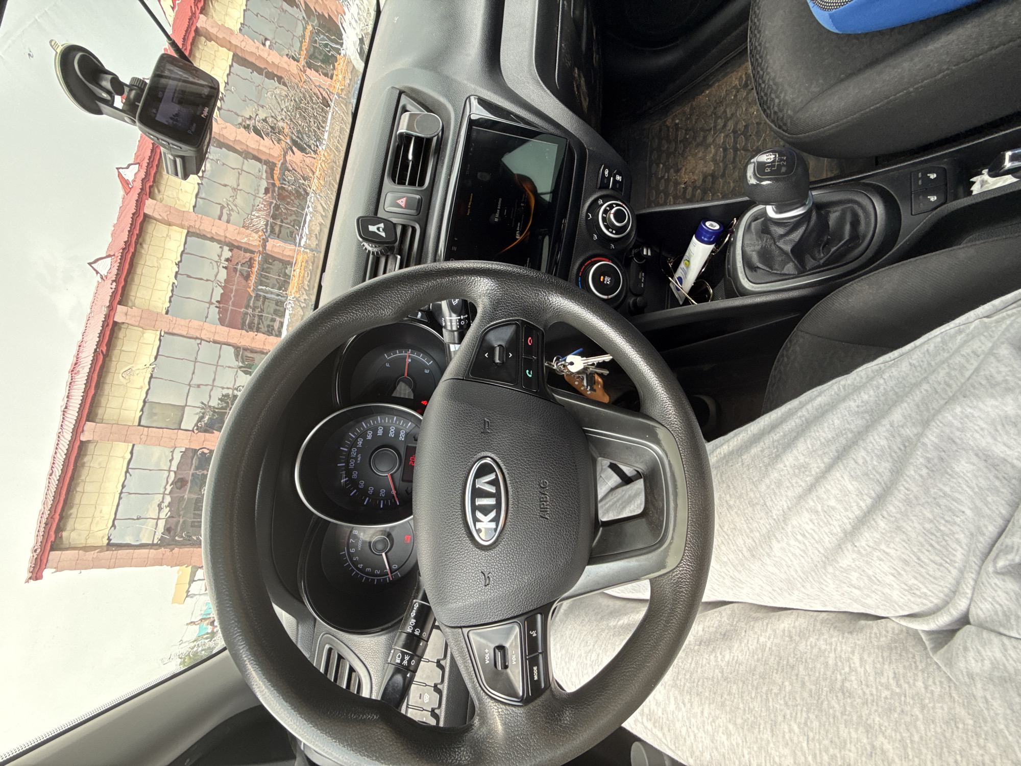 The image provides an interior view of a car, likely a Kia, from the driver's perspective. The dashboard is dominated by a steering wheel with the Kia logo prominently displayed, and a digital instrument cluster showing speed and RPM readings. To the right of the steering wheel, a touchscreen infotainment system is visible, displaying "Моя дочка" (My daughter) and some graphical elements. Below the screen are climate control knobs. A manual gear shifter with R, 1, 3, and 5 indicated is also present, suggesting a manual transmission. A dashcam is mounted on the windshield, its screen showing a reflection of a building with red and yellow facades and numerous windows. The weather appears overcast, with some water droplets on the windshield, creating distorted reflections. The car interior has dark grey plastics and black fabric seats. A person's legs, clad in light grey sweatpants, are visible in the foreground, indicating the presence of the driver or a passenger. The scene is set inside a vehicle, likely parked or driving in a location that has such buildings visible. The overall impression is a candid snapshot of a car's interior.
