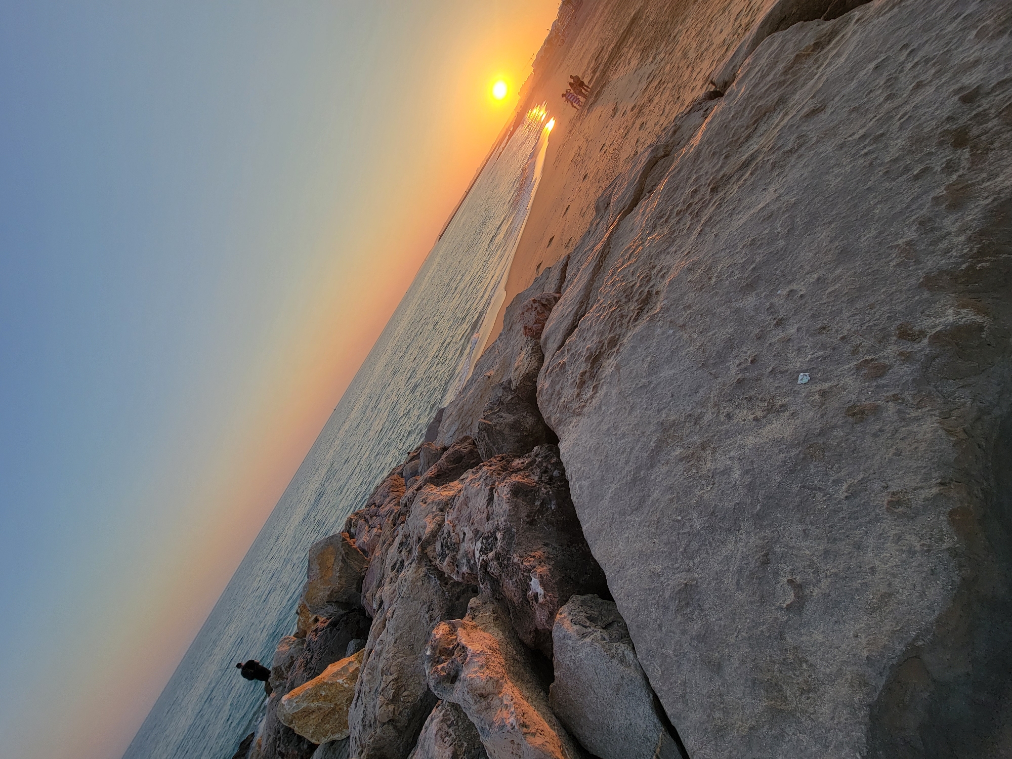 The image shows a sunset scene at the Quarteira beach in Portugal. A person is seen standing on the rocky shore, possibly fishing, with the ocean stretching out to the horizon. The sun is setting, casting a warm orange glow across the sky and reflecting on the water. Further down the beach, a small group of people are visible walking. The foreground is dominated by large, textured rocks, and the background shows the sandy beach and the calm sea. The sky transitions from a pale blue at the top to hues of orange and yellow near the horizon.