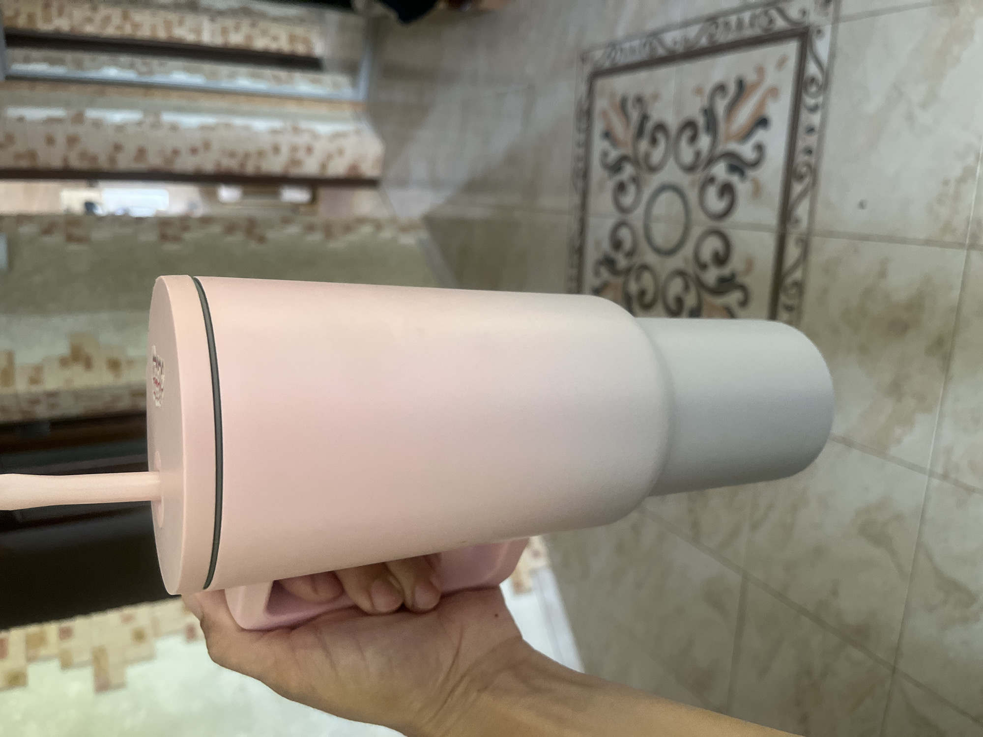 A person's hand is holding a pink and gray insulated tumbler with a pink straw. The tumbler has a handle and a black band around the top. The background shows a tiled floor and a hallway with patterned wallpaper. The image is taken indoors. The specific location in the city of Onokivtsi, Ukraine cannot be determined from the image alone.