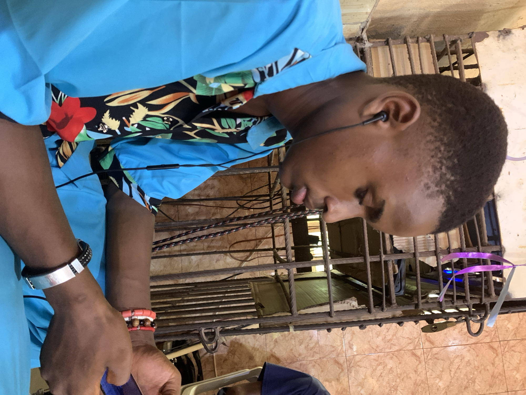 A person with dark skin and short, dark hair is wearing a light blue shirt with a floral patterned collar and has earbuds in their ears. They are standing in front of a barred structure that appears to be a window or gate. The bars are dark, and there is a padlock hanging from them. The ground is tiled with a brown and beige patterned tile. There is evidence of debris and clutter behind the bars. The setting appears to be indoors, near an exterior opening, and possibly within the city of Akpaka Forest Reserve, Nigeria. The person is looking down, possibly at something they are holding or working on. There are bracelets on their wrist.