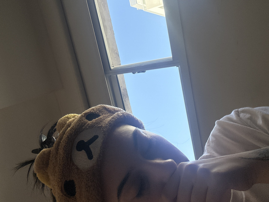 A person wears a tan, plush bear hat and a white shirt. The individual appears to be lying down, with their face angled toward the camera. A bright blue sky is visible through an open window behind them. The interior is dimly lit, creating contrast with the bright exterior. The window frame is white and appears to be partially ajar.