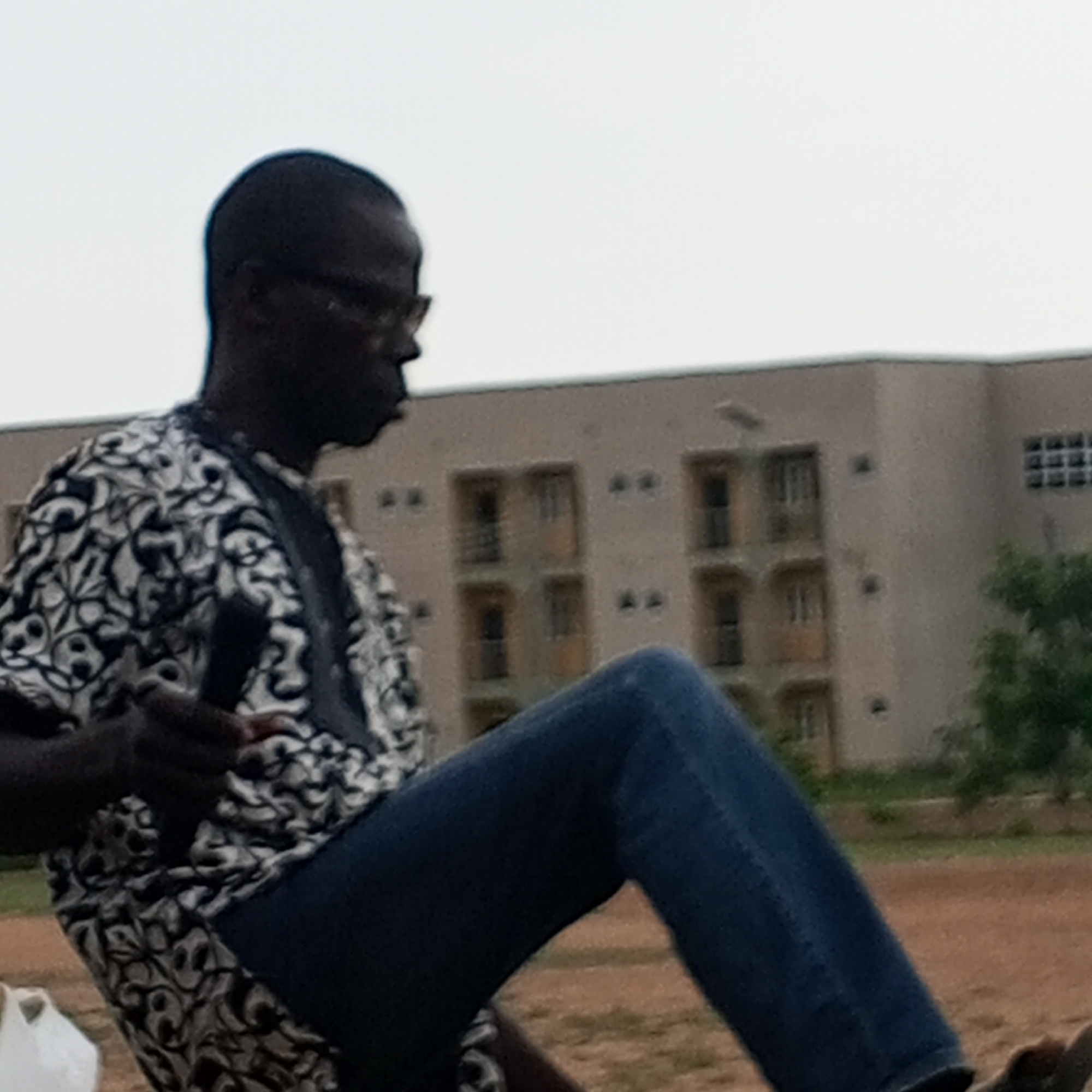 The image shows a Black man in a patterned shirt and blue jeans, sitting outdoors with his left leg raised. He is wearing glasses and holding a dark object, possibly a microphone, in his left hand. In the background, a light brown building with multiple windows is visible, along with some trees and a cloudy sky. The ground appears to be dirt. The scene is likely located in Barden Yana, Nigeria, given the style of the building and vegetation.