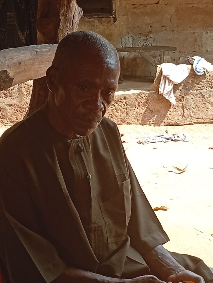 An elderly, dark-skinned man sits in the sun wearing a brown, button-down shirt with a front pocket. His head is balding, and he looks down with a serious expression. Behind him is a rough, beige stone structure with a small wooden bench. White cloths are strewn on the structure. A wooden log sits to the left of the man. The overall impression is one of age and solemnity in a rural outdoor setting.