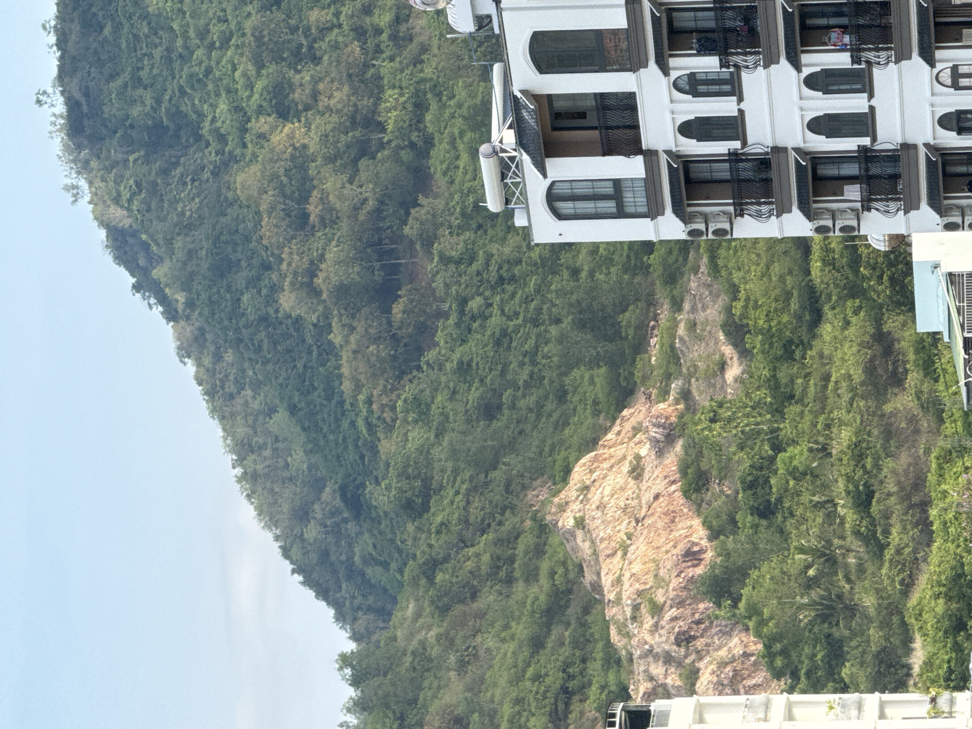 The image shows a residential building with a lush green mountain in the background. The building is white with dark balconies and arched windows. It appears to be a multi-story apartment complex. In the background, the mountain is covered in dense vegetation, with some rocky outcrops visible. The sky is clear and blue. The scene is likely in a tropical or subtropical region, given the vegetation. The cropped images provide closer views of the building's architectural details and the mountain's texture. The presence of air conditioning units on the building suggests a warm climate. The overall impression is one of a modern residential development nestled against a natural landscape.