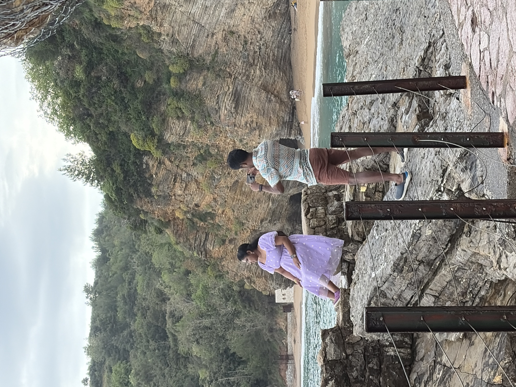 Here is a detailed description of the image:
The image captures a scenic view in Budva, Montenegro, likely taken from a high vantage point along the coastal cliffs. A man and a woman are the primary subjects. The man is taking a picture with a smartphone. He is wearing a patterned short-sleeved shirt and red shorts. The woman is wearing a light purple polka-dotted dress. They are both on the rocky cliffs overlooking a beach and the Adriatic Sea. The cliffs are interspersed with trees and vegetation. A protective railing runs along the edge of the cliffs. The beach below is populated with several people. The overall weather is partly cloudy, and the location gives a panoramic view of the coast in Budva.