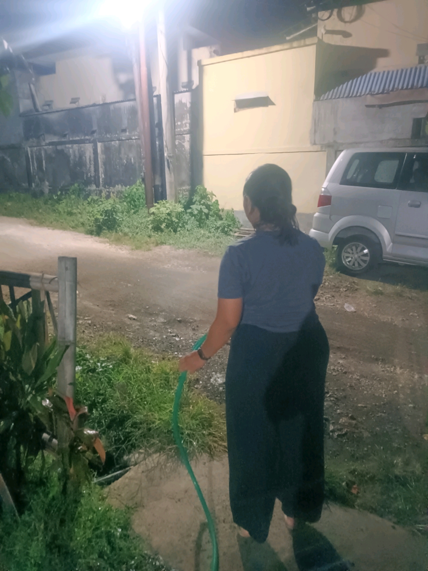 A woman, seen from behind, stands on a dirt and grass path, holding a green garden hose. She is wearing a dark blue t-shirt and dark pants. Her dark hair is pulled back. To her left, a metal fence post and some green foliage are visible. In the background, a light-colored car is parked on the right side of the path. Behind the car, a yellow building with a striped awning can be seen. To the left of the car and behind the fence, a dark, possibly metal, gate is present, adjacent to a light-colored building. The scene appears to be outdoors at night, illuminated by a bright light source above and to the left of the frame. The ground is damp, suggesting recent watering or rain. The location is identified as the city of Tabanan, Indonesia.