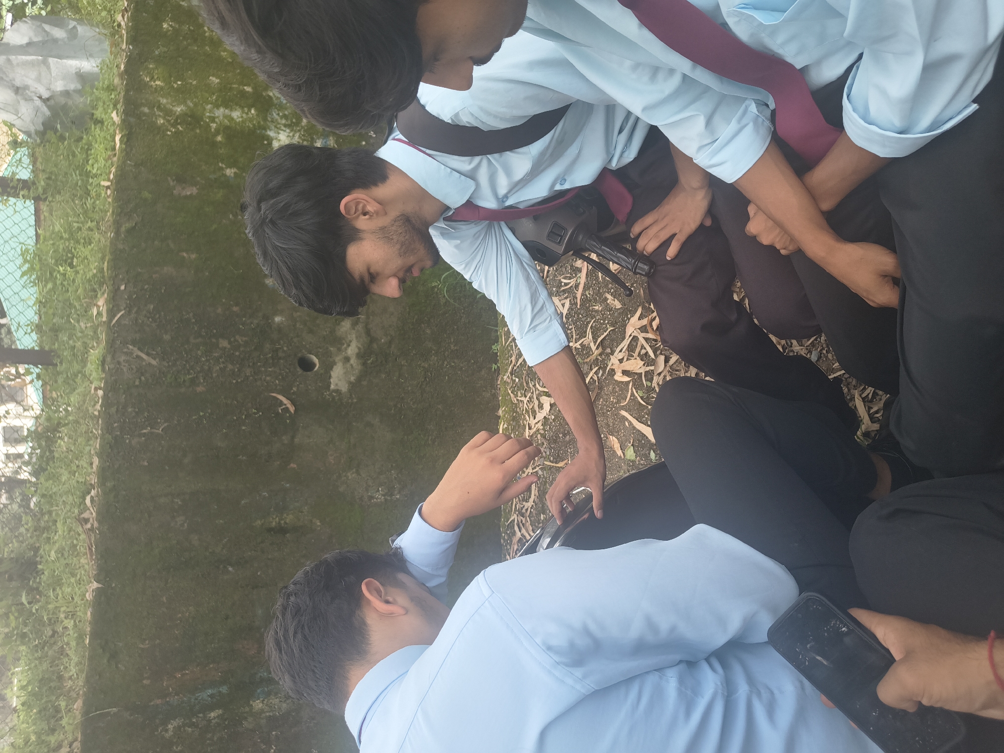 Three young men in light blue collared shirts and dark pants are sitting outdoors on the ground, possibly on a scooter, near a moss-covered concrete wall. One man on the left is leaning his head on his arm, looking down at a smartphone he is holding. Another man in the center is looking down at the phone with his left hand resting on his knee and his right hand touching the scooter. A third man on the right is partially visible, looking down and to his left. The ground is covered with dry leaves and dirt. In the background, there is greenery and a fence. The overall lighting suggests it is daytime. The image appears to have been taken in Dharamshala, India, likely in a university or school setting given the attire.