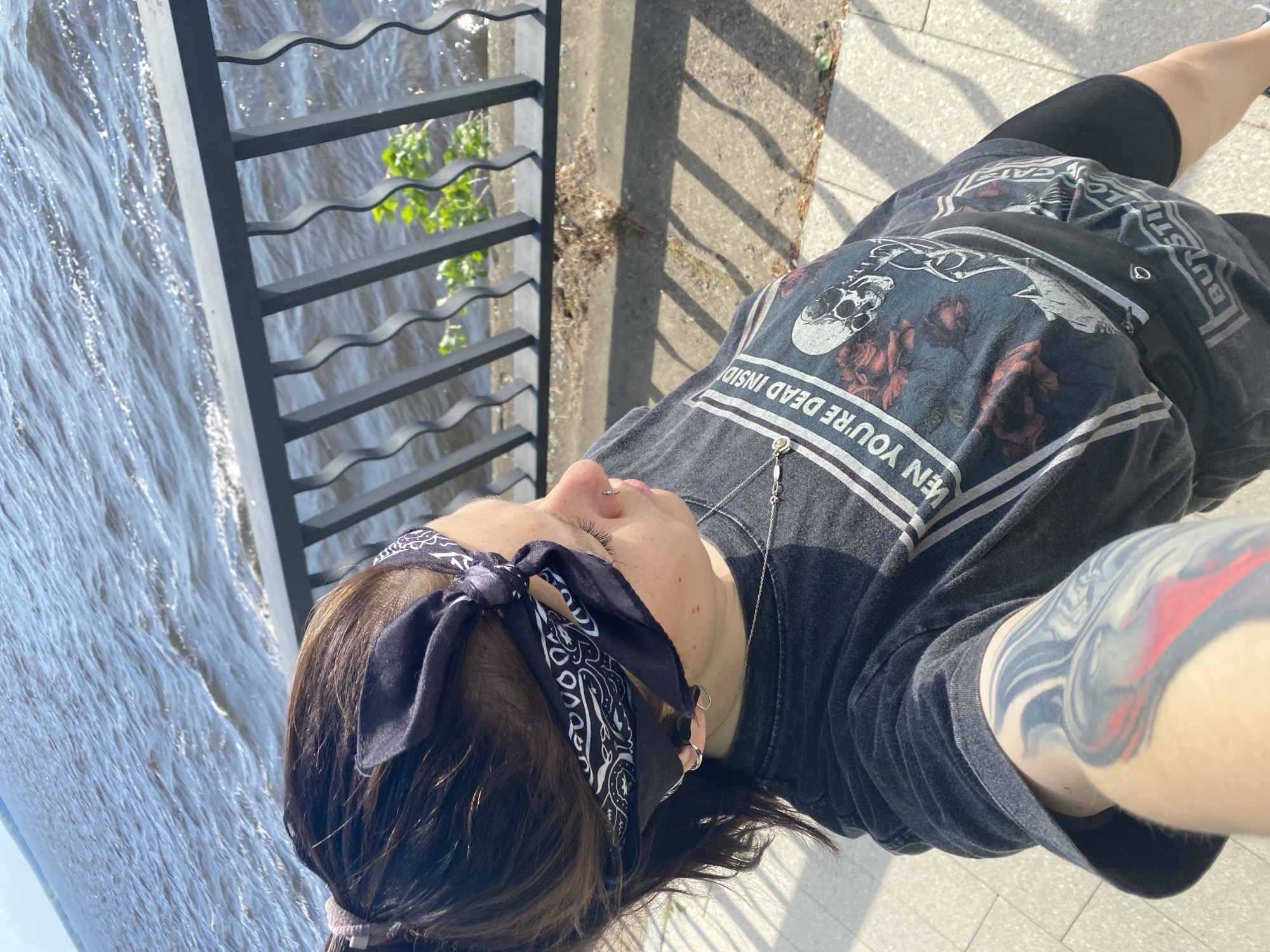 A person is standing on a sidewalk in the city of Chaikovsky, Russia, next to a wrought iron fence. The person is wearing a dark t-shirt with a skull and floral design, black shorts, and a bandana. The fence overlooks a body of water. The person has tattoos on their arm. The sidewalk is made of light-colored pavers. Shadows from the fence are cast on the sidewalk.