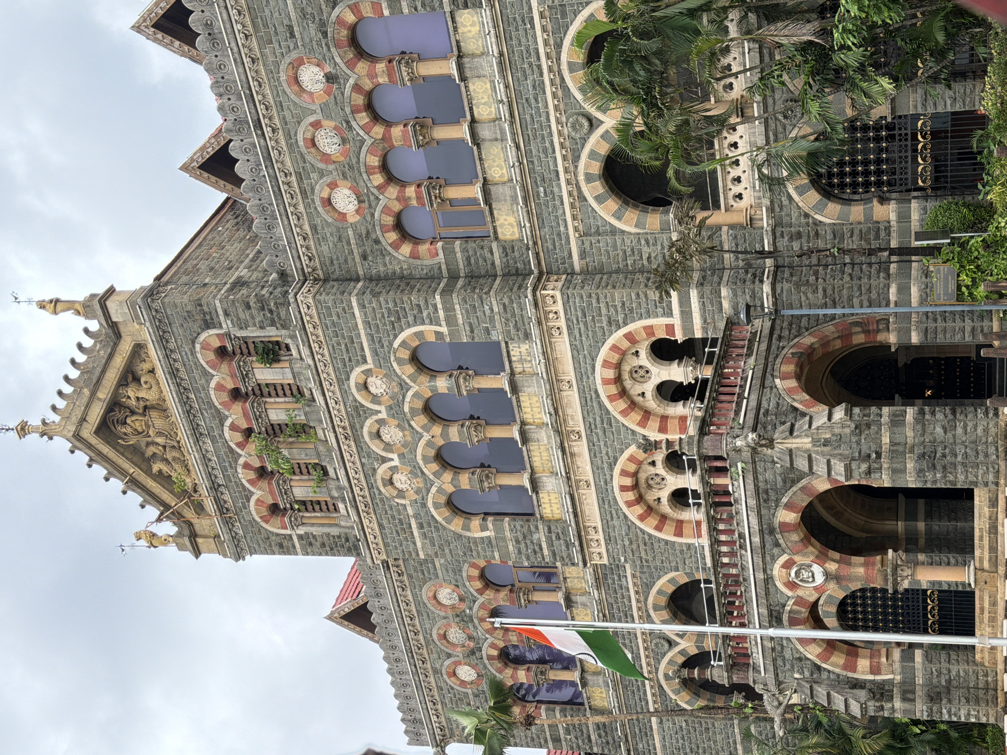 The image captures the exterior of the Chhatrapati Shivaji Maharaj Terminus (CSMT), formerly Victoria Terminus, a historic railway station in Mumbai, India. The architectural style is a fusion of Victorian Gothic Revival and Indian architectural elements. The building is constructed with grey basalt stone and features intricate carvings, arched windows with colored glass, and a grand facade. An Indian flag is prominently displayed on a flagpole in the foreground, to the left of the building. Palm trees and other lush greenery surround the structure, suggesting a tropical or sub-tropical environment. The sky is overcast, indicating a cloudy day. The image focuses on the upper stories of the building, showcasing its detailed ornamentation, including statues and decorative friezes. There are no people visible in the image.