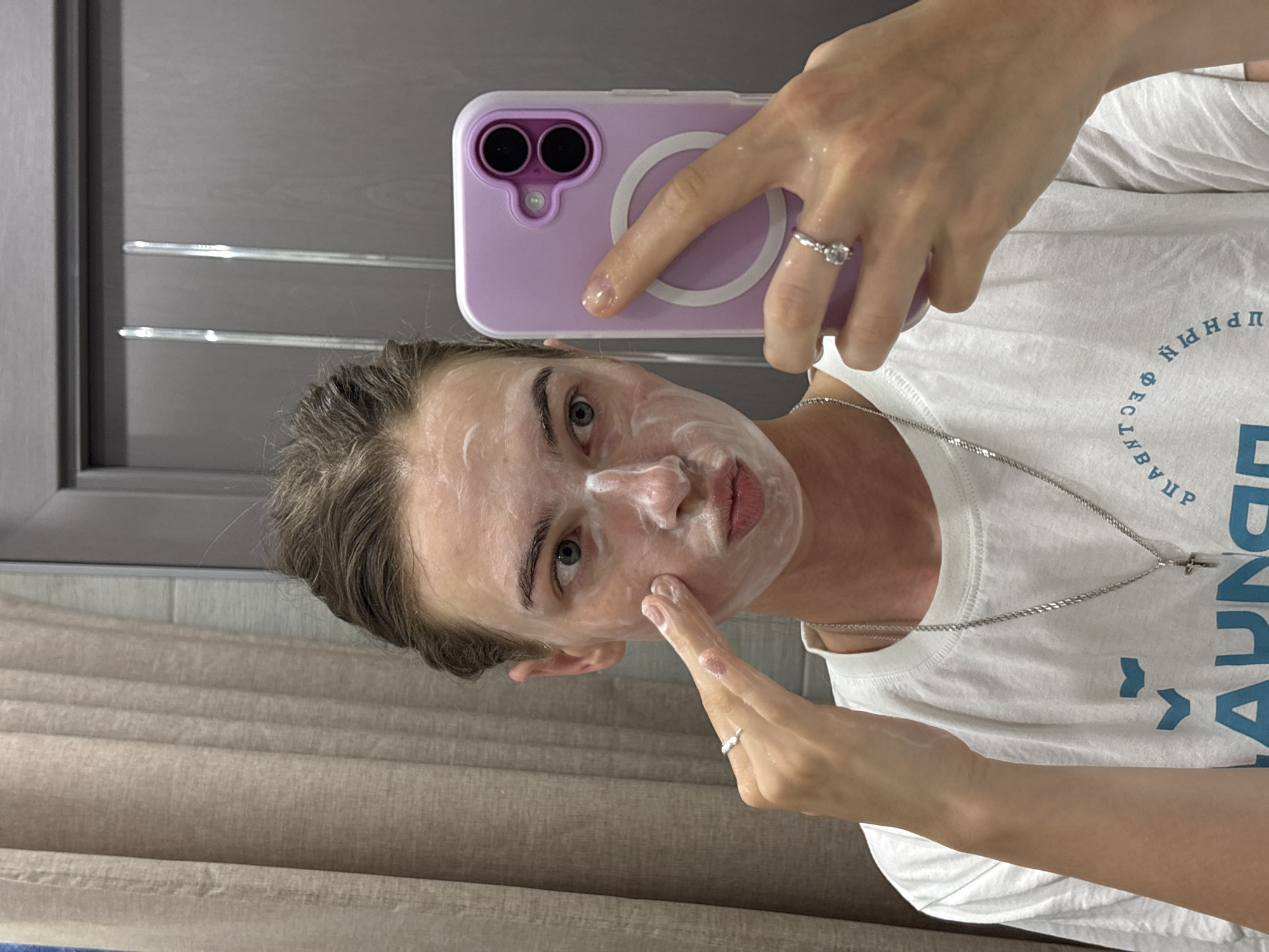 A young woman with her hair pulled back is taking a selfie in a mirror. She has white cream or mask on her face. She is wearing a white t-shirt with a silver necklace. Her left hand is touching her face, and her right hand is holding a light purple phone case. The background shows a beige curtain and a grey door. This scene could be in a bathroom in the city of Perm, Russia.