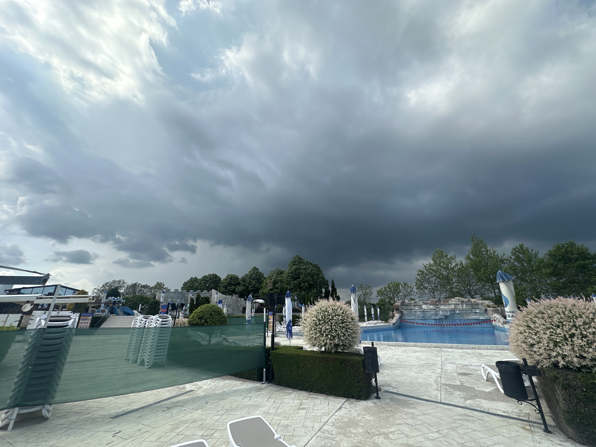 The image depicts a scene at a pool in the city of Ravda, Bulgaria, under a stormy sky. The pool area, with blue water, features stacked white lounge chairs and closed blue and white umbrellas. Manicured bushes are scattered around the perimeter, and a green screen partially obscures a building in the background. The overcast sky is dominated by dark, heavy clouds.