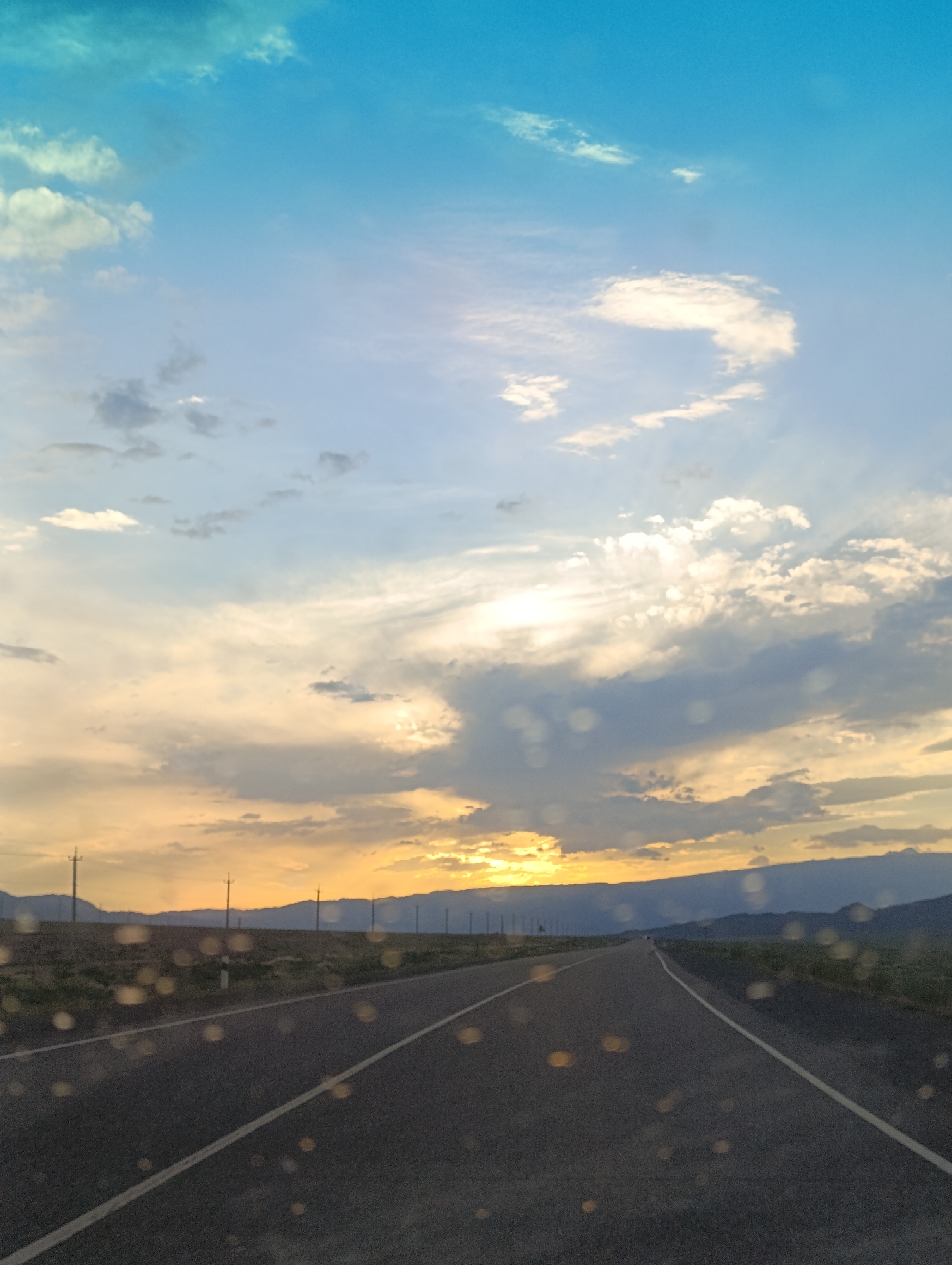 A road stretches into the distance under a dramatic sky at sunset, possibly in the vicinity of Orto Tokoy, Kyrgyzstan. The asphalt road has a white dashed line marking the lane. On either side of the road, there are sparse fields with scattered utility poles. In the background, a range of dark mountains is visible against the horizon. The sky is a spectacle of colors, transitioning from a bright, almost teal blue at the top to hues of yellow and orange where the sun is setting. Clouds are scattered throughout, some appearing fluffy and white, while others are darker and more textured, catching the golden light of the sunset. The foreground of the image is slightly blurred, and there are numerous circular, out-of-focus highlights, possibly indicating raindrops or water droplets on a windshield or lens. There are no people or animals visible in the scene.