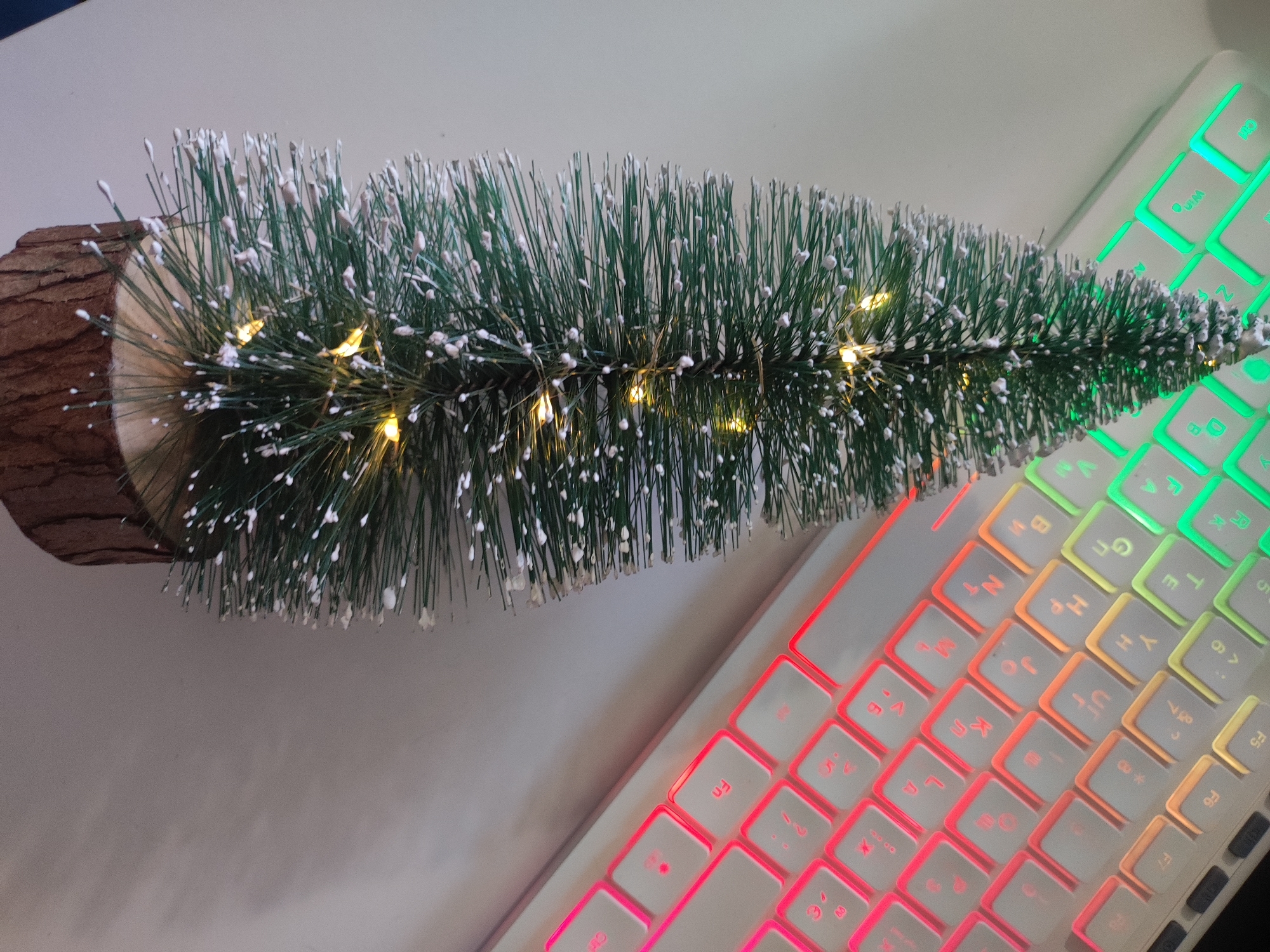 The image displays a miniature artificial Christmas tree with a log base, adorned with white flocking and small, warm-toned LED lights. The tree is positioned diagonally across a white keyboard with customizable RGB backlighting. The keyboard keys are illuminated in various colors, transitioning from green on the upper right to red on the lower left. The background is a plain, light-colored surface. There are no people or animals visible in the scene, nor any indication of the city of Cherkasy, Ukraine. The image appears to be a still life arrangement, possibly for decorative or festive purposes.