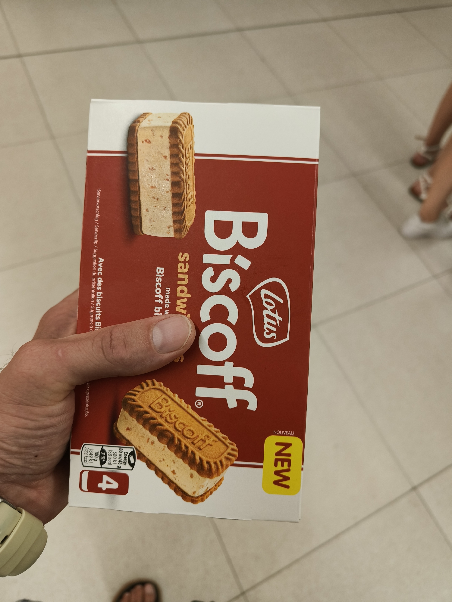 The image shows a hand holding a Biscoff sandwich cookie box. The box is predominantly red with white text and a picture of the cookie. The cookie appears to be a sandwich of two Biscoff cookies with a cream filling. There are nutritional facts and a "NEW" sticker on the box. The background is a light-colored tiled floor, suggesting an indoor environment. The image appears to have been taken in Gandia, Spain, given the text visible on the box.