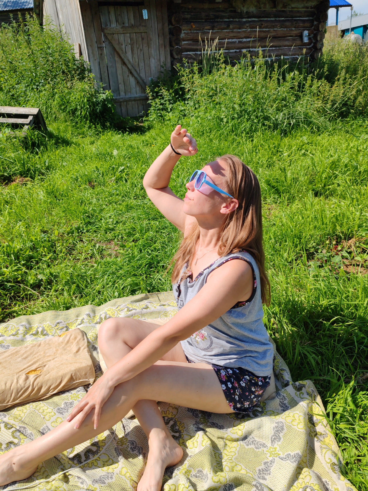 In a rural outdoor setting, likely in or around the city of Chukhlom, Russia, a woman sits on a blanket on a grassy lawn. She is positioned in a relaxed, seated pose with her legs crossed and her left arm raised, hand shading her eyes as she looks upwards. She is wearing a gray sleeveless top with a floral pattern on the neckline and dark shorts with a floral print. Blue-rimmed sunglasses rest on her nose. Beside her on the blanket is a light brown pillow. Behind the woman, a wooden structure, possibly a shed or outbuilding, stands amidst tall green grass and vegetation. The overall scene suggests a sunny day, possibly for leisure or relaxation outdoors.