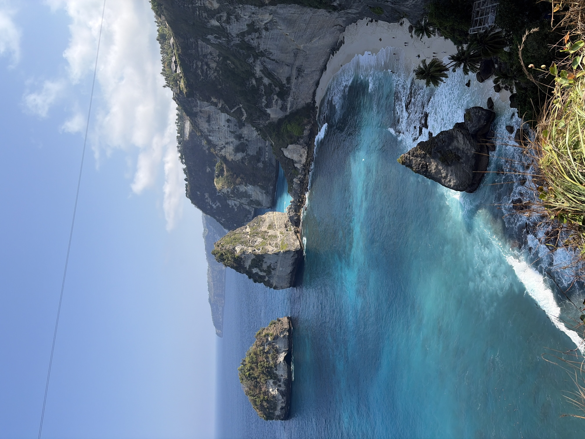 This is a high-angle, full shot of a coastal landscape in Nusa Penida, Indonesia, showcasing Diamond Beach. The image features a dramatic cliff face on the right, descending to a pristine white sand beach. The ocean displays a gradient of vibrant turquoise and deep blue hues, with gentle waves breaking along the shore. Several large rock formations, including distinctive diamond-shaped ones, are scattered in the water and along the coastline. The cliffs are covered in lush green vegetation and trees. A few people are visible on the beach, appearing as small figures walking or standing. In the distance, more rocky outcroppings and the coastline of Nusa Penida are visible under a clear blue sky dotted with wispy clouds. A thin, dark line, likely a power line, stretches across the top of the frame.