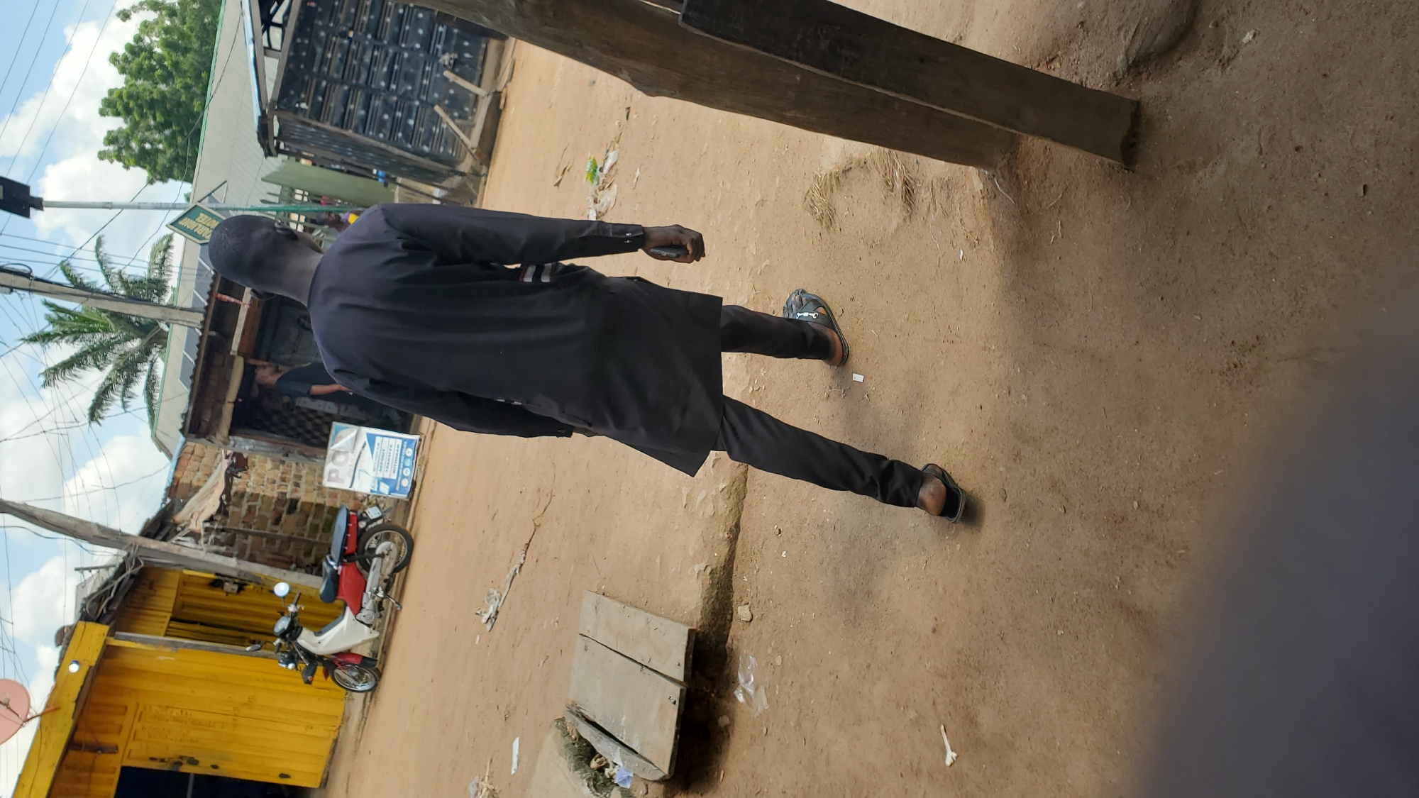 A man walks away from the camera, wearing a black tunic and trousers and black sandals. He is on a dirt road in what appears to be a market or commercial area in Jalingo, Nigeria. To his left, a yellow corrugated metal building is visible with a red and white motorcycle parked in front of it. Behind the man, a row of shops with metal shutters is present. The sky is cloudy.