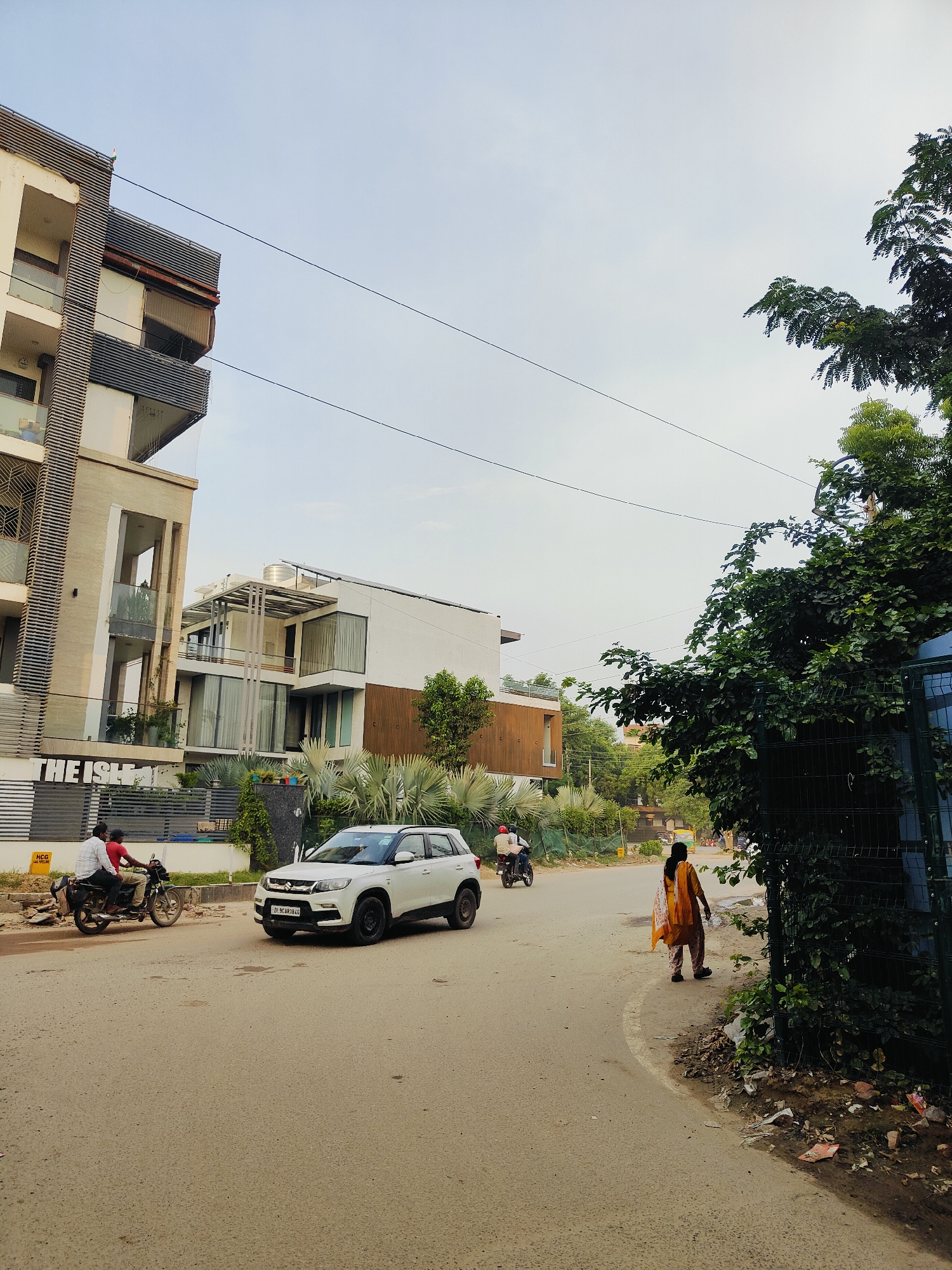 In Gurgaon, India, an urban street scene unfolds under a pale sky. On the left, a modern, multi-story building with a contemporary facade stands tall. Its design features a mix of white and beige materials, with prominent horizontal louvers and glass balconies. In front of this building, a black metal fence runs along the property line.
On the right side of the street, a woman wearing a yellow and orange patterned outfit walks away from the camera towards the right edge of the frame. She is on the sidewalk, which is littered with debris. Adjacent to the sidewalk is a dense cluster of green trees and foliage, some of which are growing through a dark green chain-link fence.
The road in the foreground is paved and appears to be moderately trafficked. A white SUV, identified by a license plate reading "DL 9CAR 2845", is parked or driving on the right side of the road. Further down the road, a motorcycle with two occupants is visible, heading towards the left. The general environment suggests a developing or established residential area within the city, characterized by modern architecture and lush greenery.