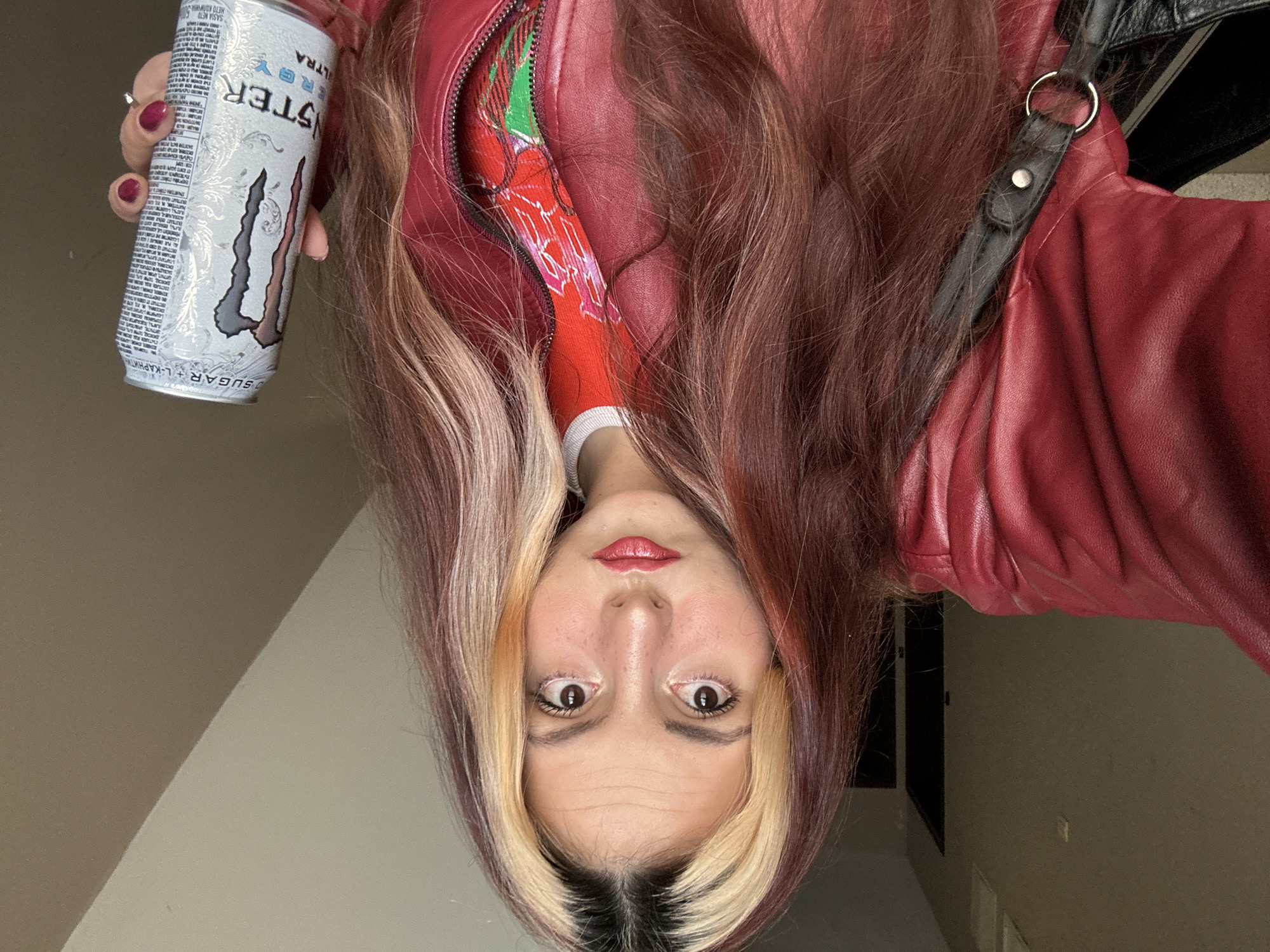 A woman is holding a can of Monster Energy Ultra drink. She is wearing a red leather jacket with a red and white patterned shirt underneath. Her hair is long, with blonde streaks in the front and red highlights throughout. The photo appears to be taken indoors. The location cannot be determined from the image.