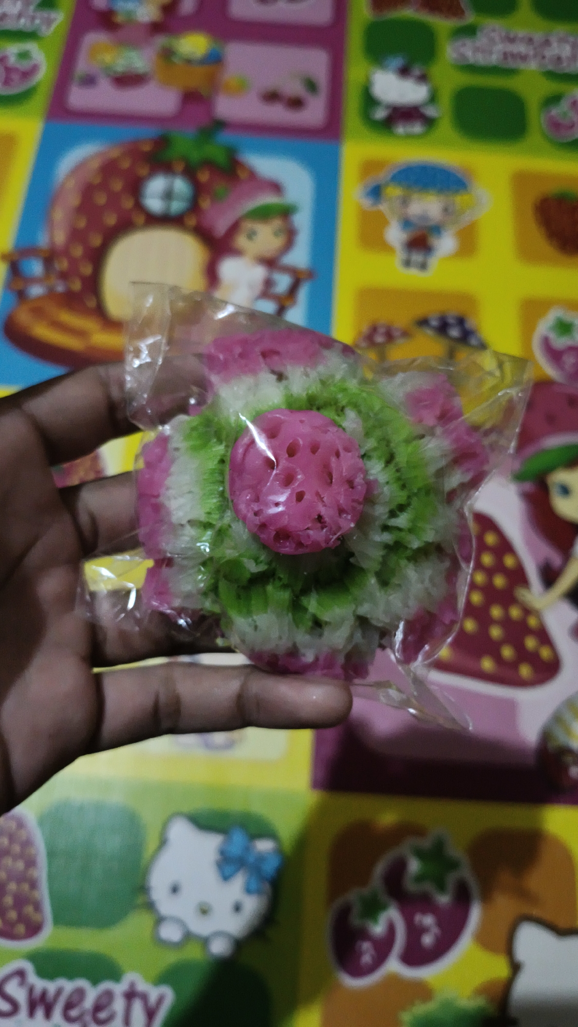 A hand holds a pink and green flower-shaped candy wrapped in clear plastic. The candy has a textured pink center. In the blurred background, there is a colorful mat with cartoon characters, including strawberries, Hello Kitty, and other figures. The mat appears to be spread on a surface in Kerep Kidul, Indonesia.