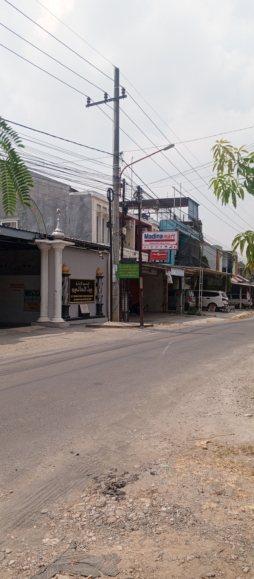 This is a street-level, eye-level, medium-distance, full-shot photograph of a street in Gulun, Indonesia. The scene depicts a street with a rough asphalt surface. On the left side of the street, there is a building with a gray facade and a white decorative pillar topped with a silver dome. Adjacent to this building, there is a sign with Arabic script. Further down the street, there are several commercial buildings. One building has a red and white sign that reads "Madina mart" and displays various logos. Another sign beneath it shows "Jon & Parcel". Several cars are parked along the street, including a white SUV. Overhead, a complex network of electrical wires and poles runs across the sky, which is overcast. To the left and right edges of the frame, there are glimpses of green foliage from trees. The road itself shows tire marks and patches of gravel and debris.