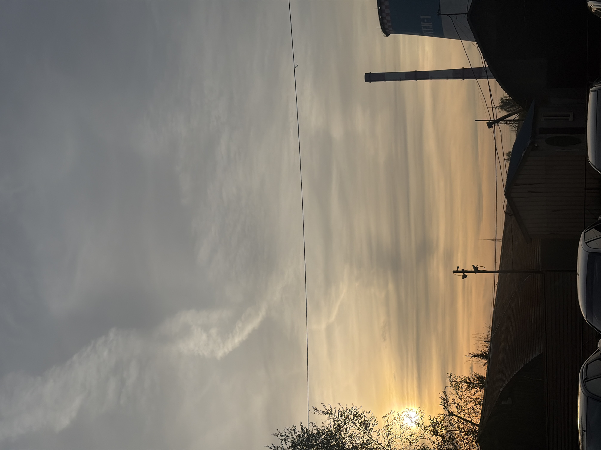 The image shows an industrial landscape at sunset. Visible are a tall cooling tower labeled "TЭЦ-1" (TEC-1), a smokestack, and a building. A utility pole and electrical wires are also present. The sky is filled with clouds and the sun is setting. Parts of cars are visible. The scene is most likely located in Saint Petersburg, Russia, where many combined heat and power plants are located.