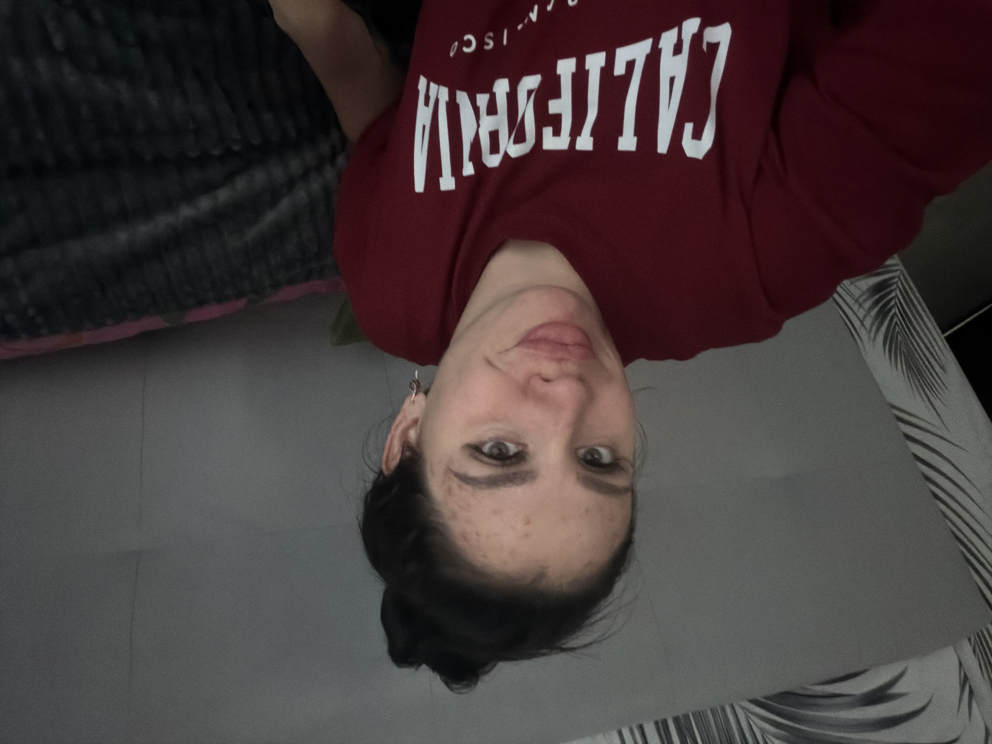 The image shows a young woman with dark hair pulled up in a bun, wearing a maroon t-shirt with "CALIFORNIA" written in white on the front. She has fair skin with freckles and is looking at the camera with a slight smile. She is wearing hoop earrings. The background is out of focus and appears to be an indoor setting, possibly a bedroom, with a grey headboard and a patterned throw blanket. There is no indication of the city of Фоки, Russia.