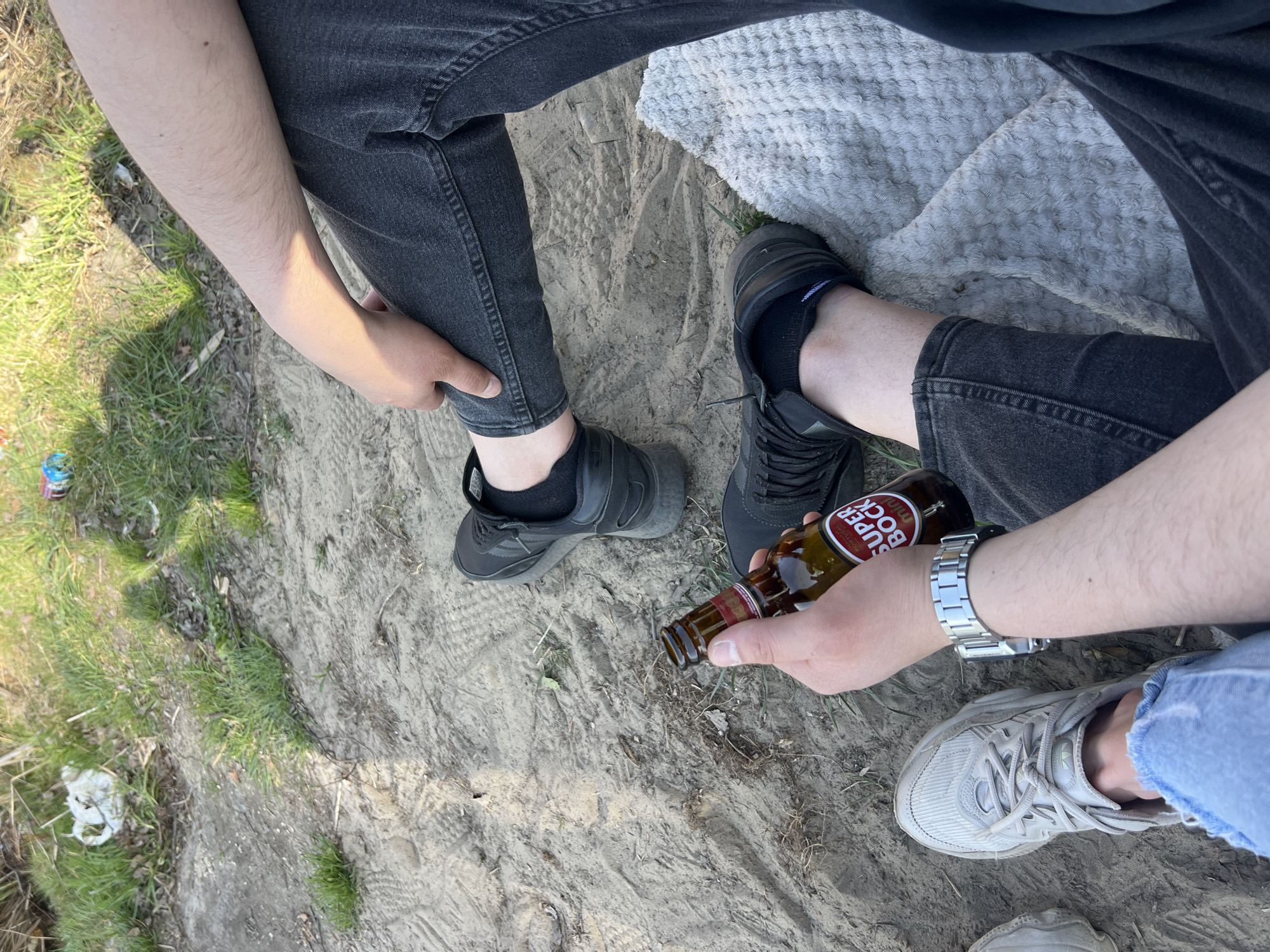 The image depicts a ground-level view of two individuals sitting outdoors, likely in Ciemne, Poland, though specific location details are not evident. The surface is a mix of light-colored dirt and sparse grass. The individuals are wearing dark-colored pants, and their footwear consists of dark sneakers and light-colored sneakers. One person is holding a brown glass bottle labeled "Super Bock." A silver wristwatch is visible on the wrist of the person holding the bottle. A white cloth or blanket is partially visible in the background. Litter is visible on the ground.