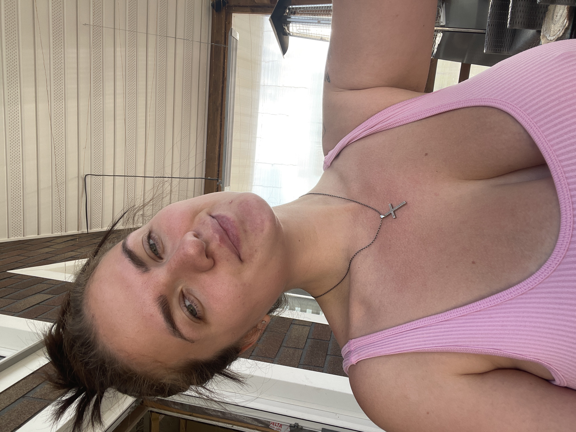 The image features a woman posing for a close-up shot, angled from below, looking towards the camera. She has fair skin, green eyes, and dark brown hair pulled back, with some strands escaping around her face. She wears a light pink, ribbed tank top and a silver cross pendant necklace. Her expression is neutral and direct.
The setting appears to be outdoors on a bright day, possibly on an elevated structure like a balcony, patio, or rooftop, aligning with the provided location context of Мосметаллоконструкция Онт, Russia. In the background, to the left, are vertical cream-colored panels or blinds, with thin wires running down them. To the right, a bright, hazy outdoor scene is visible through a clear panel or window. Below and to the left of the woman's head, brown roof shingles with white trim are visible, suggesting a roofline or building structure. Parts of a dark metal frame or furniture are also present in the upper right.
Notable details include the bright lighting, suggesting sunshine. Text visible on a label in the bottom left corner reads "DELTA MULTI BAND" and "ULTRA". The woman's head is slightly tilted.