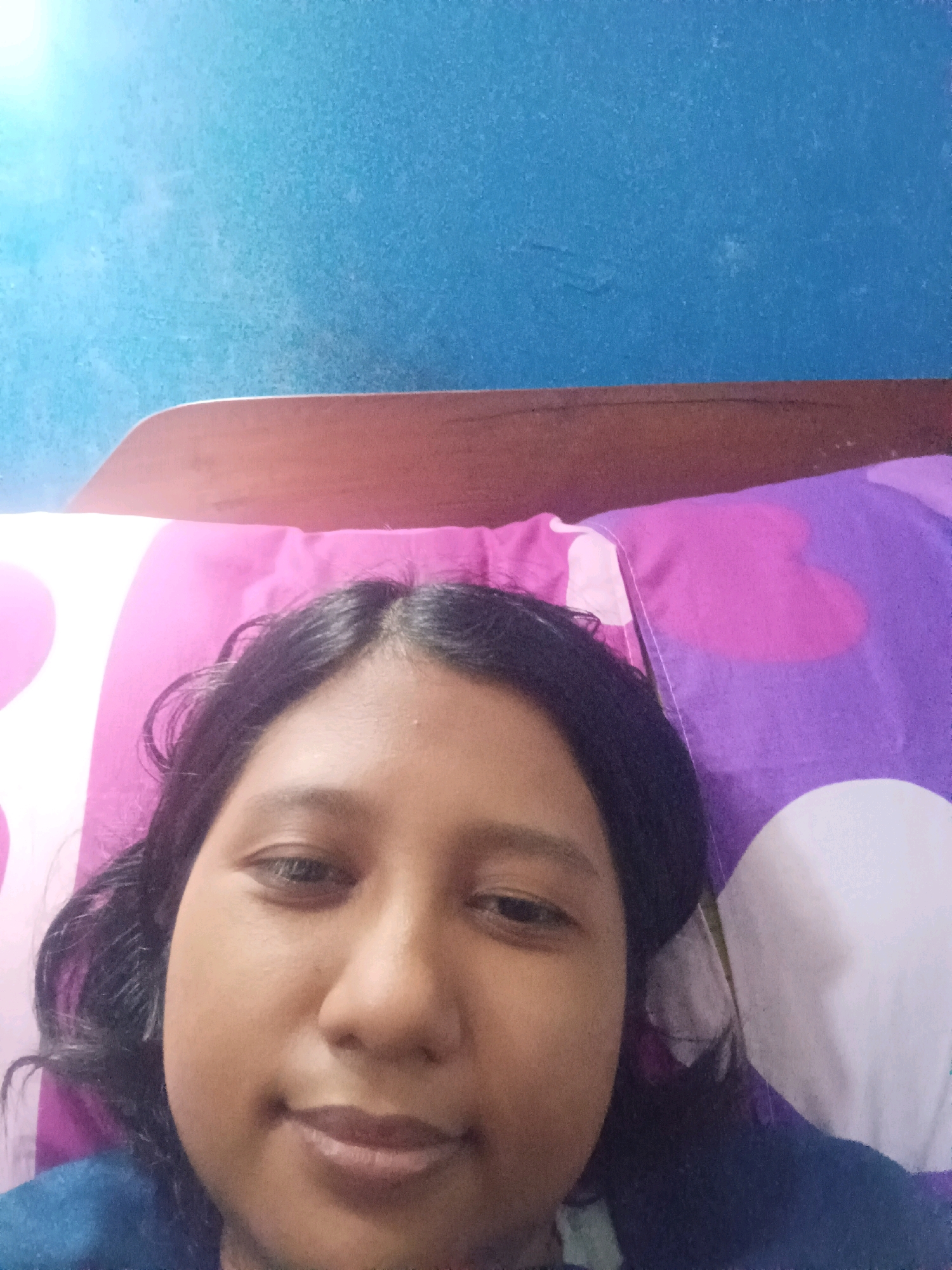 This is a close-up, slightly blurry, selfie of a young woman lying in bed. She has dark hair and is wearing a blue shirt. She is looking directly at the camera with a slight smile. The background shows a blue wall and a pillow with a pink and purple heart pattern. The image appears to have been taken indoors. There are no specific visual cues that would indicate the location within the city of Canga'an, Indonesia.