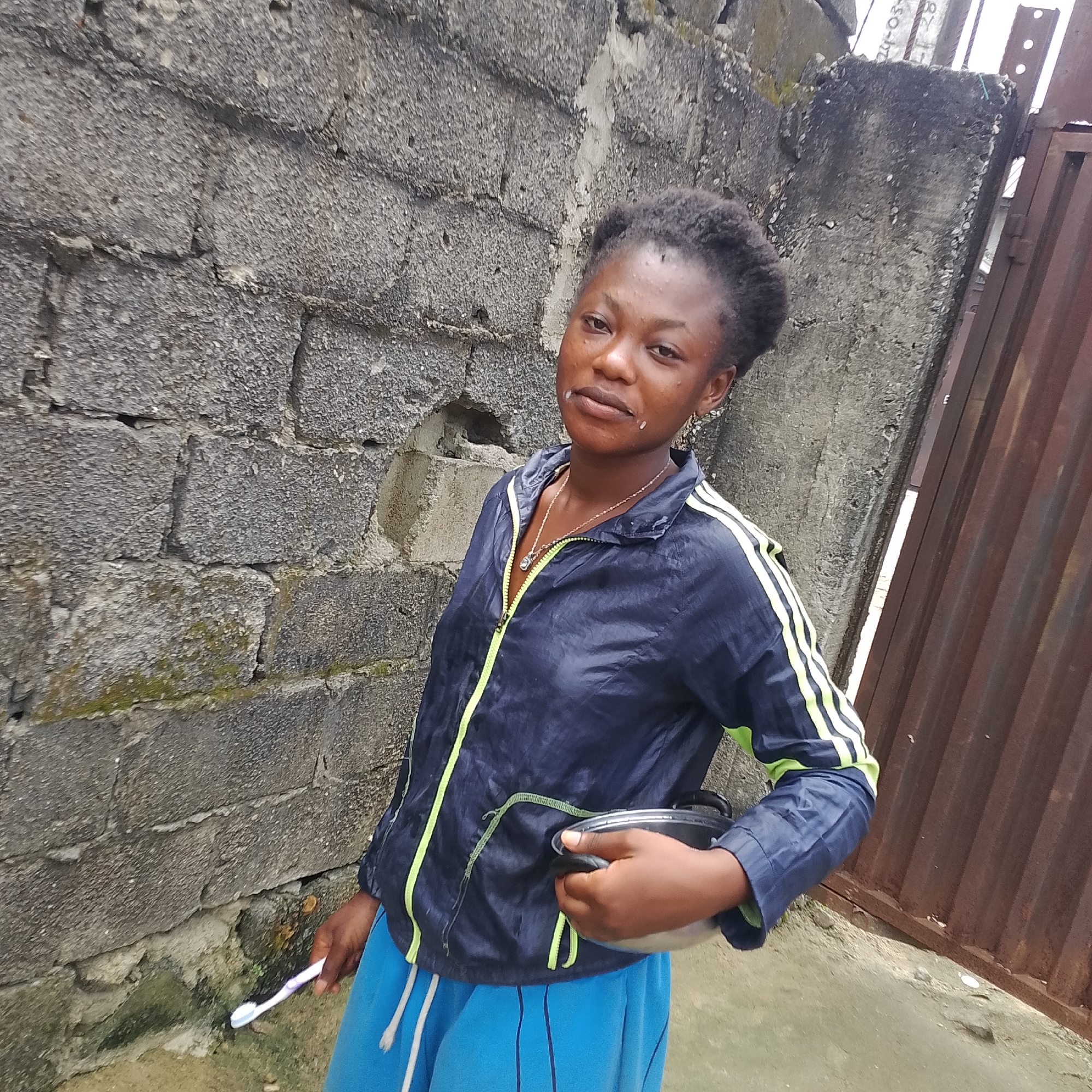 A young Black woman, appearing to be in her late teens or early twenties, stands outdoors in front of a rough, gray cinder block wall and a rusted corrugated metal fence. She is wearing a dark blue athletic jacket with bright yellow trim and blue athletic pants. Her hair is pulled back in a dark bun. She is holding a small, silver metal bowl in her right hand and a toothbrush in her left. There is a substance that appears to be toothpaste or foam around her mouth. The ground is a mix of dirt and concrete. The background suggests an urban or semi-urban environment. The overall lighting indicates it is daytime.