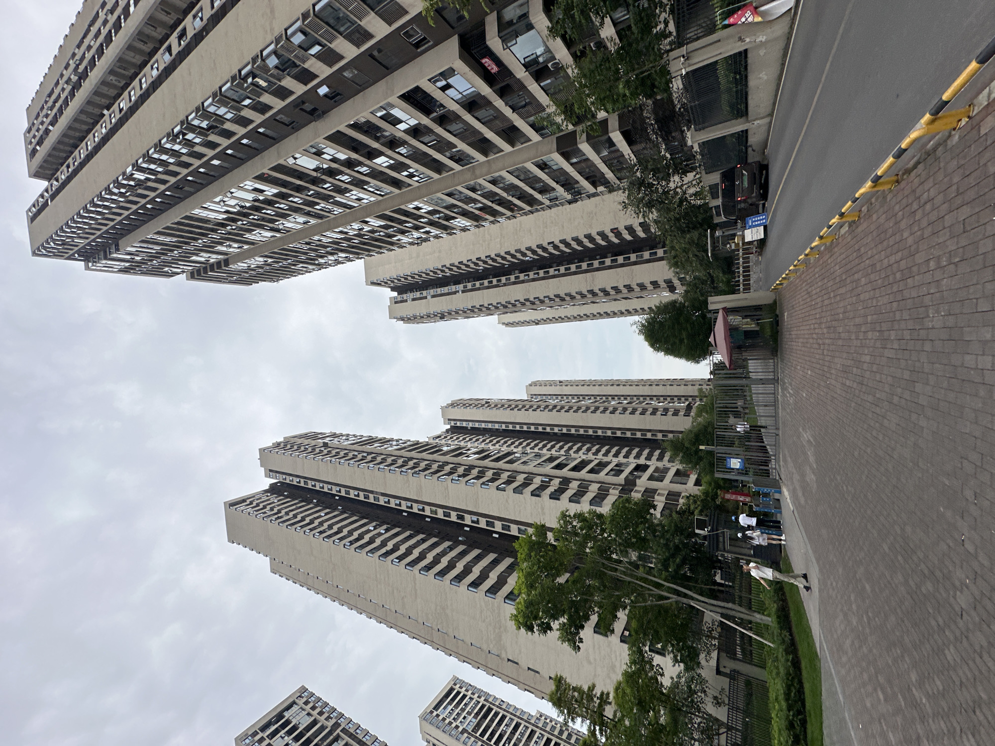 The image captures a dense urban residential scene in the Chongqing High-tech Zone, China, featuring numerous high-rise apartment buildings under an overcast sky. The photograph is oriented vertically, with tall, multi-story buildings dominating the left and center, rising towards a muted grey sky.
The buildings share a common architectural style: light beige concrete or stucco facades with dark, deeply recessed windows and balconies arranged in repetitive patterns. Their sheer height and number suggest a modern, densely populated area. Many windows reflect the cloudy sky or appear dark.
At ground level, to the right of the buildings, a paved road runs parallel to a brick-paved sidewalk. A continuous yellow and black striped barrier separates the roadway from the pedestrian path. Further down the street, a black SUV is parked. Adjacent to the parked vehicle, a blue rectangular sign is visible, displaying Chinese characters which include "智谷" (Zhi Gu, meaning "Smart Valley") and "二期" (Phase Two), identifying a specific area within the high-tech zone.
Several trees with green foliage are interspersed between the buildings and the ground level, adding patches of natural color to the concrete environment. On the sidewalk, a few individuals are present. One person, dressed in light-colored clothing, walks away from the camera. Closer to a red awning or umbrella, a small group of people, some wearing white shirts, are gathered, possibly near an outdoor recreational or exercise area that is partially obscured by fencing and greenery.
The weather appears to be overcast, with diffused daylight, indicating daytime but without direct sunlight. The overall scene depicts a typical residential streetscape within a modern high-tech urban development.