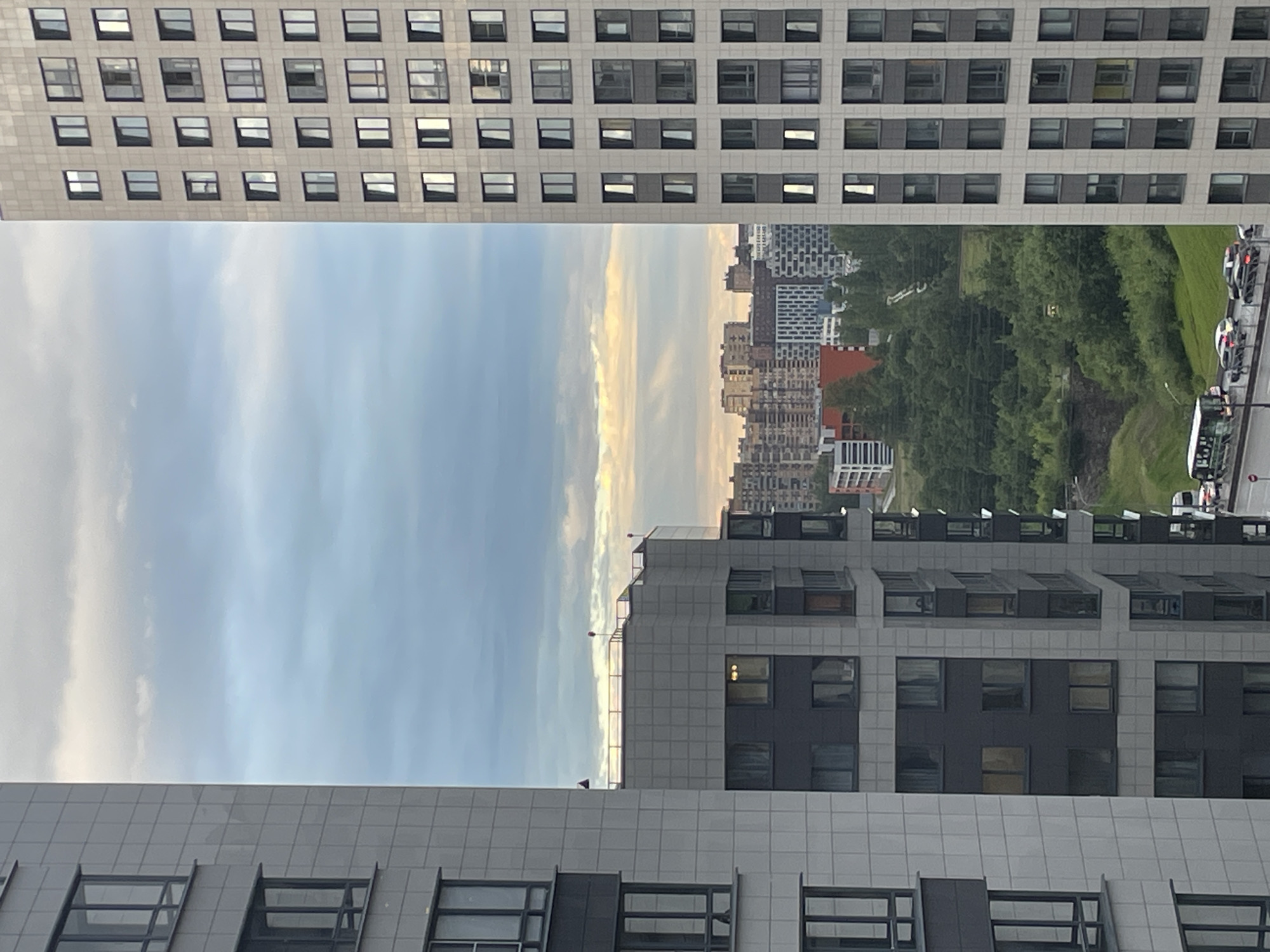 This media file displays a view of modern apartment buildings with numerous windows, set against a backdrop of a cloudy sky and a cityscape. The buildings are constructed with light-colored facade panels and dark-colored window frames. Some windows reflect the sky, while others reveal interior lights, suggesting it is either dusk or dawn. In the distance, more buildings of varying heights and architectural styles are visible, interspersed with lush green trees. A road with vehicles, including a white bus, is also present on the right side of the image. The overall scene appears to be an urban environment, possibly a residential complex. The lighting suggests it is late afternoon or early evening, with the sky exhibiting soft, diffused light and some areas of warmer hues, possibly from the setting sun. No people are clearly discernible in the image. No text is visible.